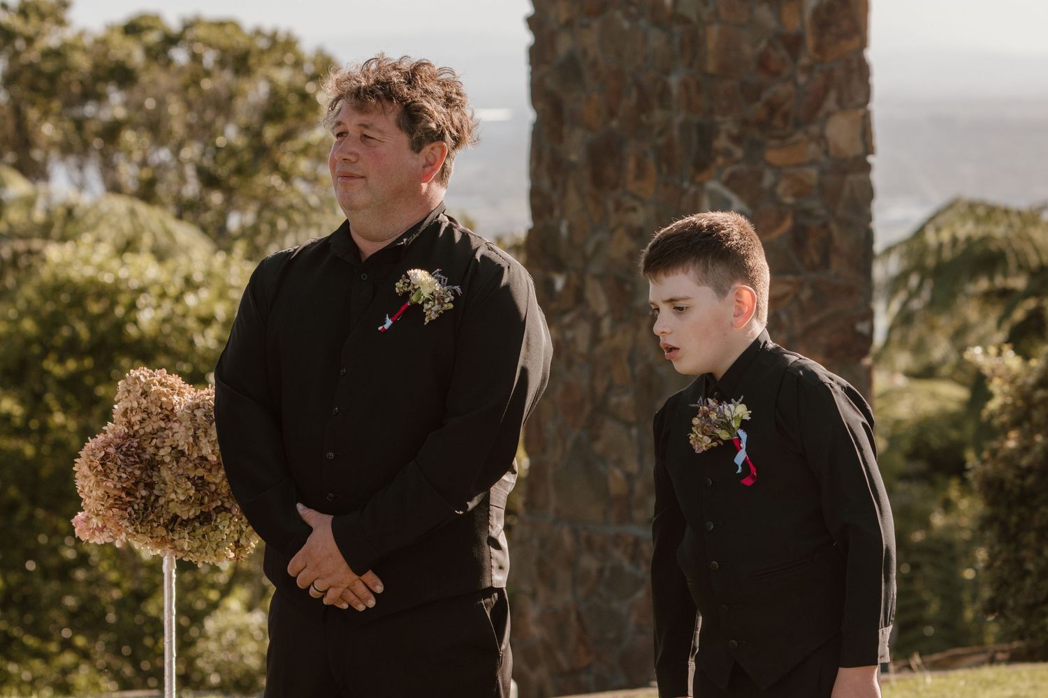 A stylish group wearing black suits stand together during a sunny outdoor wedding ceremony at a rustic stone venue.