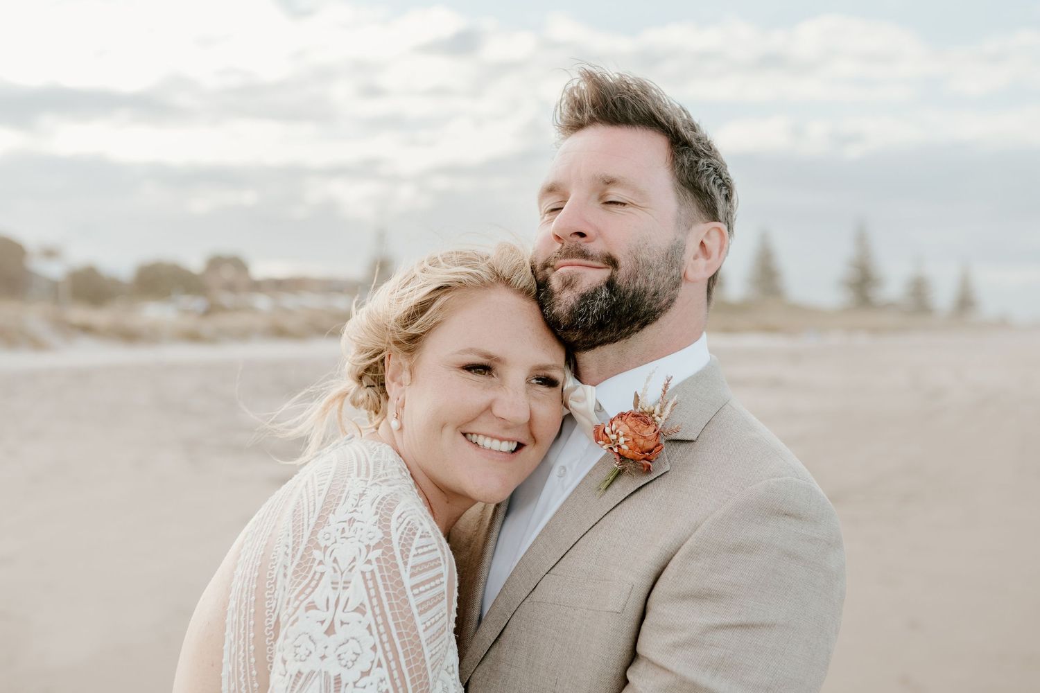 Wedding couple sharing an intimate moment on a beach with a coral boutonniere and beige suit on a cloudy day.