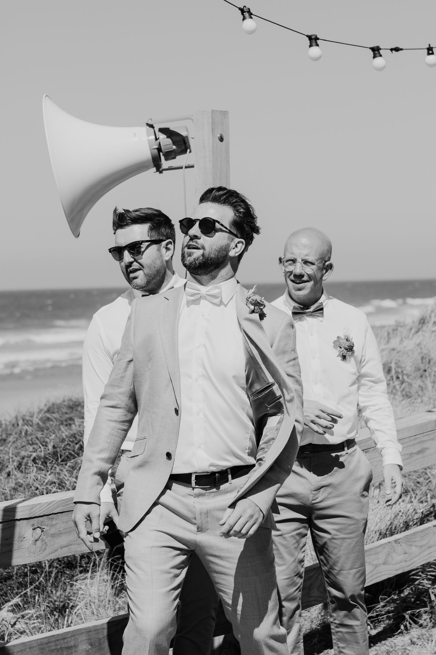 Black and white photo of groomsmen in sunglasses at beach wedding with string lights and megaphone in casual attire.