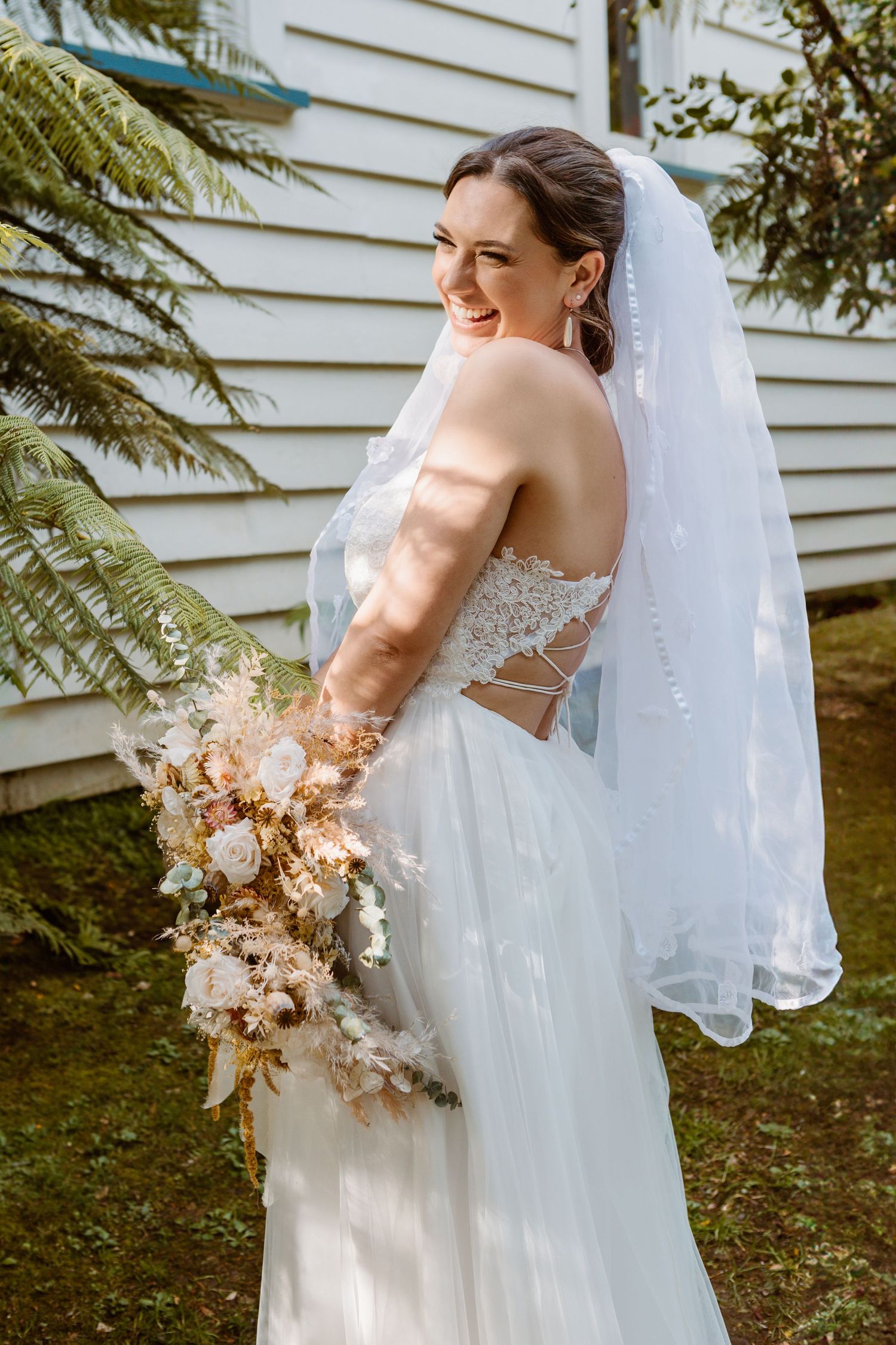 A bride in a lace white wedding dress and veil holding a dried floral bouquet against a white building exterior.