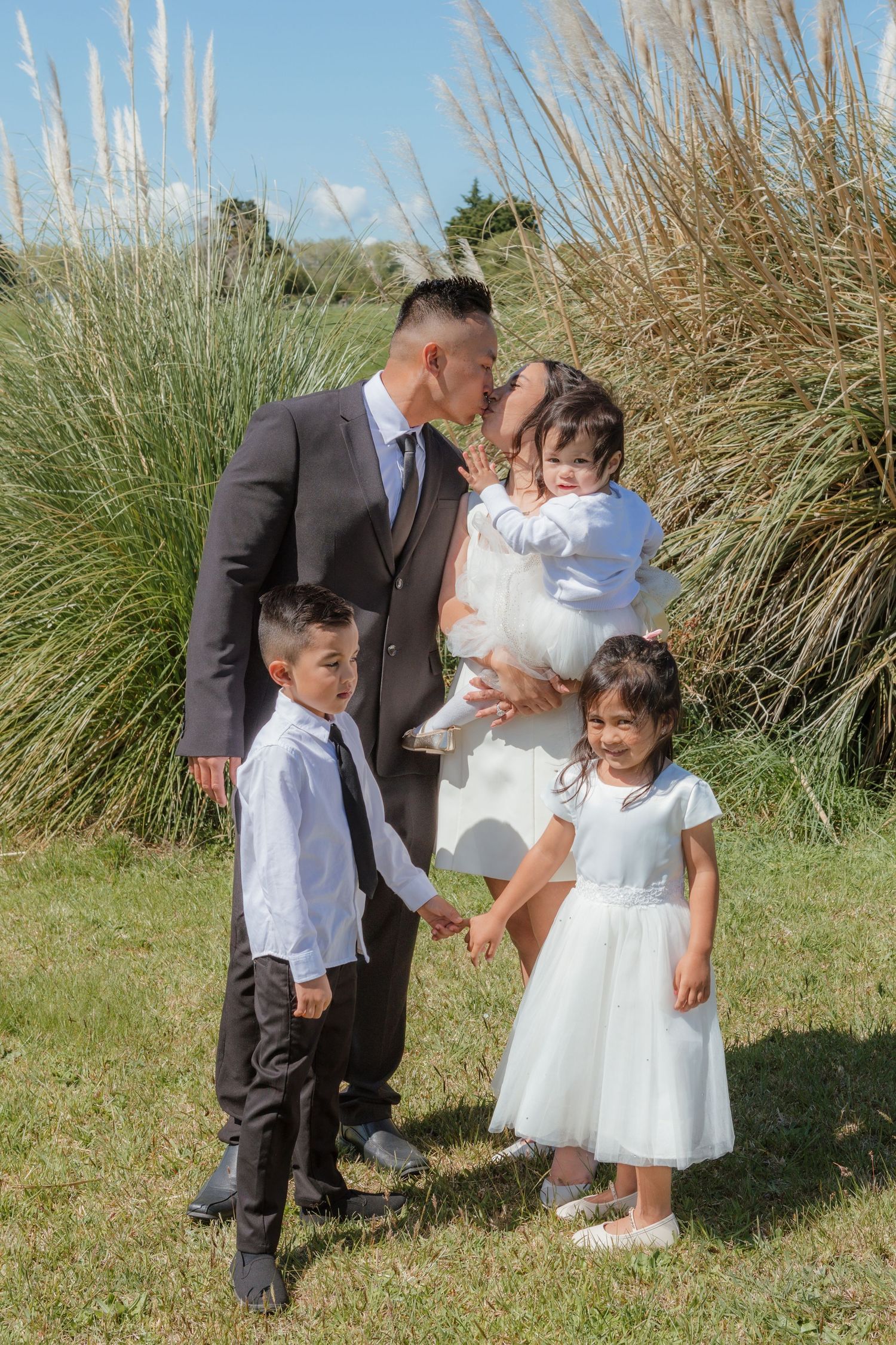 Young children in formal wedding attire playing together in tall beach grass on a sunny day.