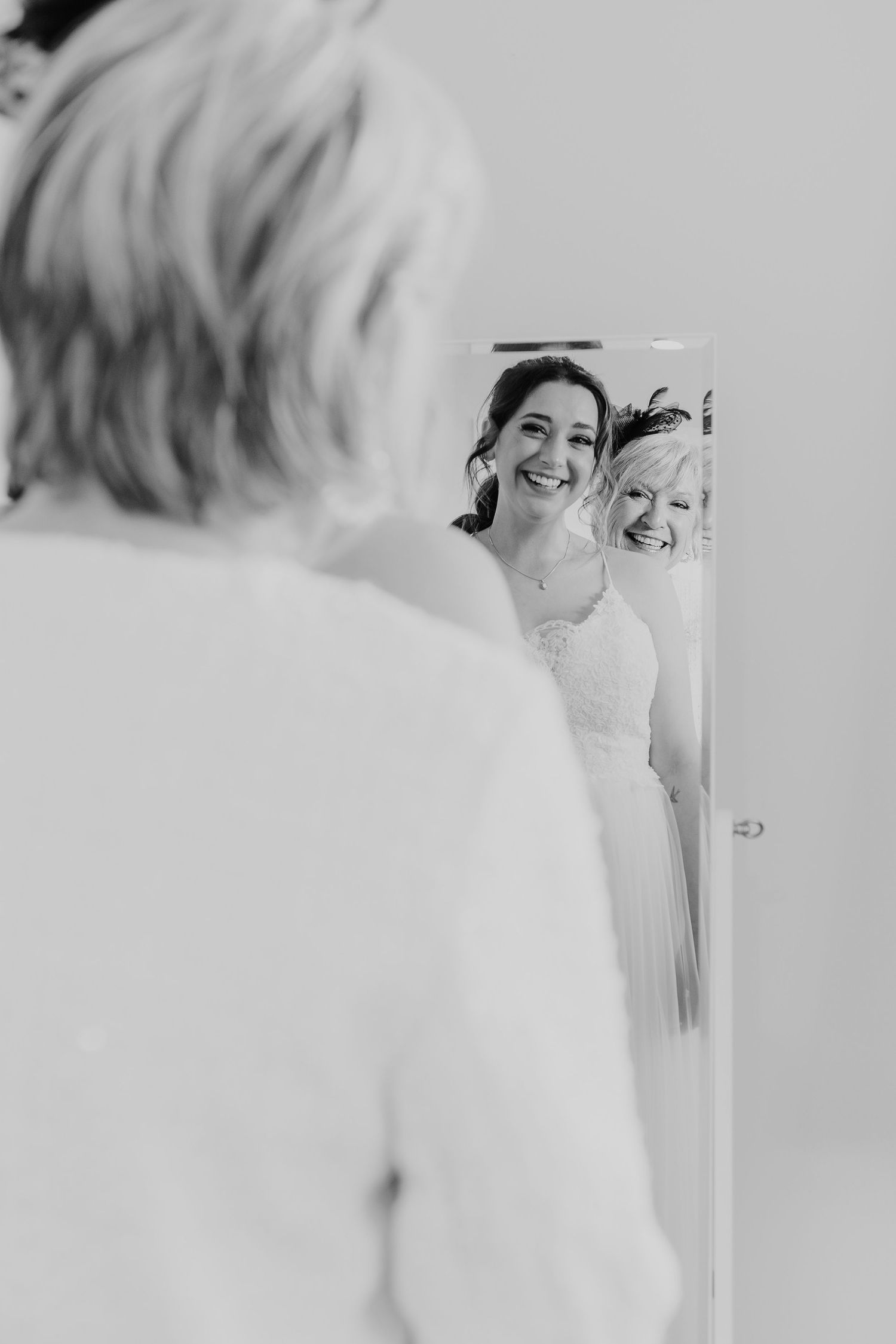 A joyful bride looks at her reflection in a mirror during wedding preparations, captured in black and white.