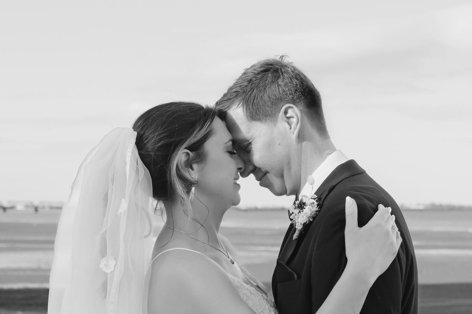 A romantic black and white wedding portrait with a couple embracing as their foreheads touch, with a flowing veil visible.