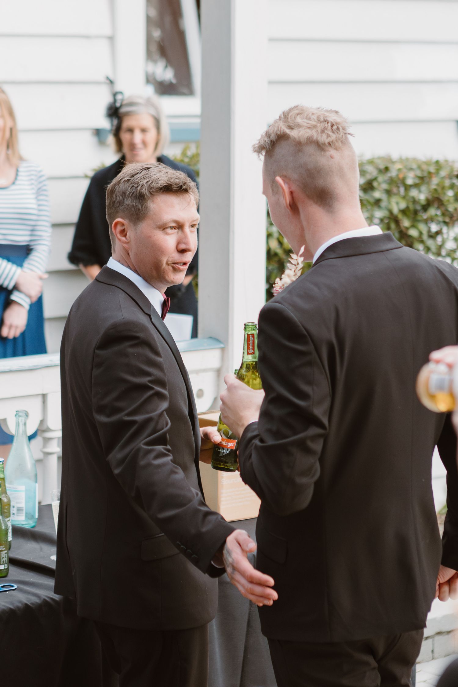 Two well-dressed men in black suits having a conversation while holding drinks at an outdoor event.