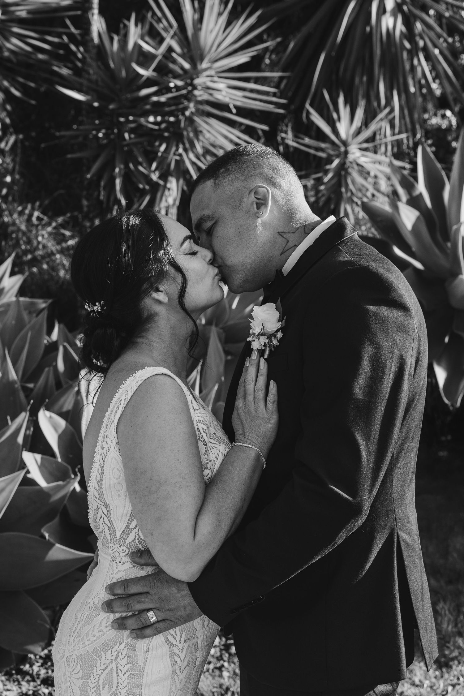 Black and white wedding photo show newlyweds embracing outdoors with tropical foliage in background. Bride and groom portraits in Tauranga