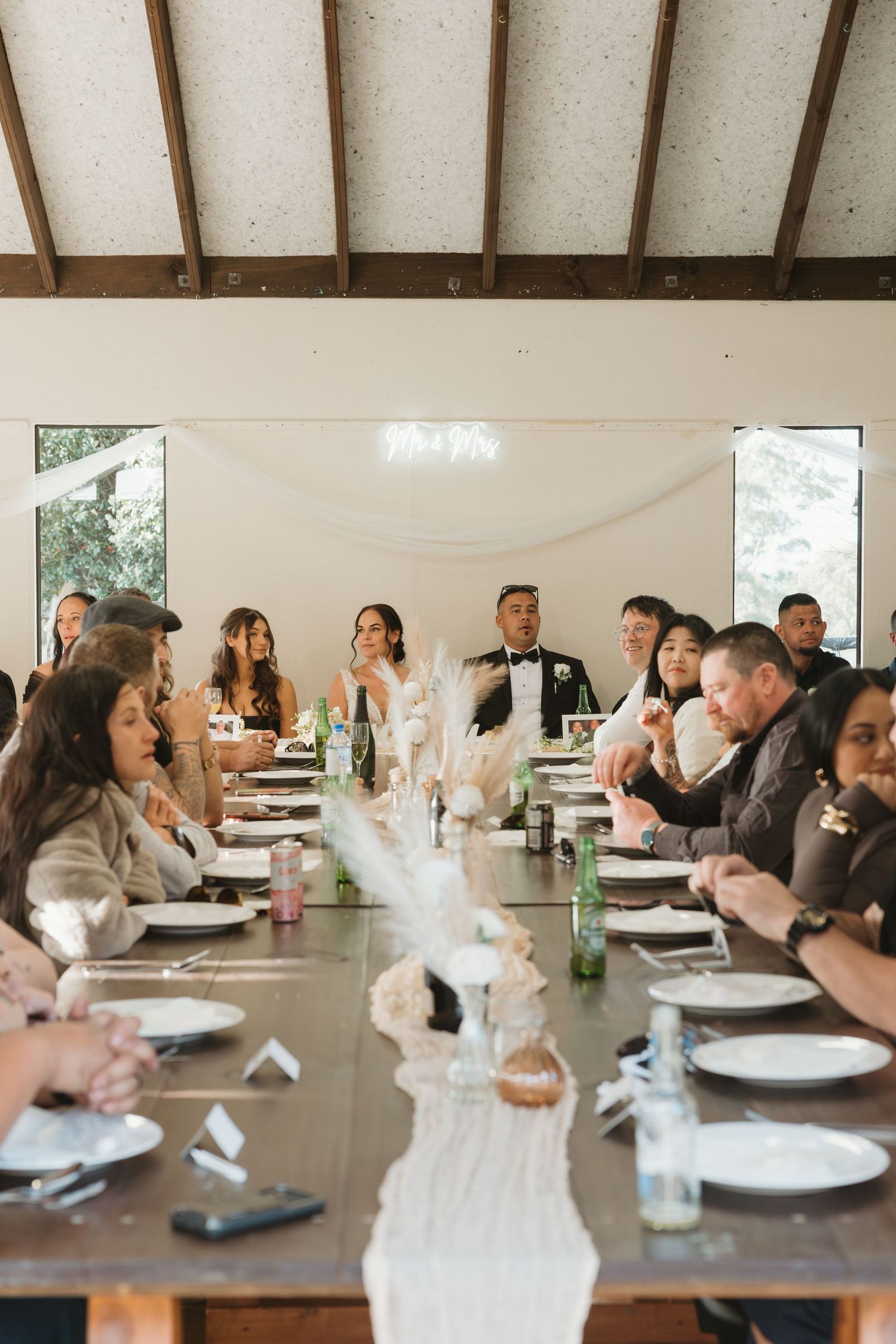 Wedding guests seated at long reception tables with rustic decor and white place settings in a bright venue.