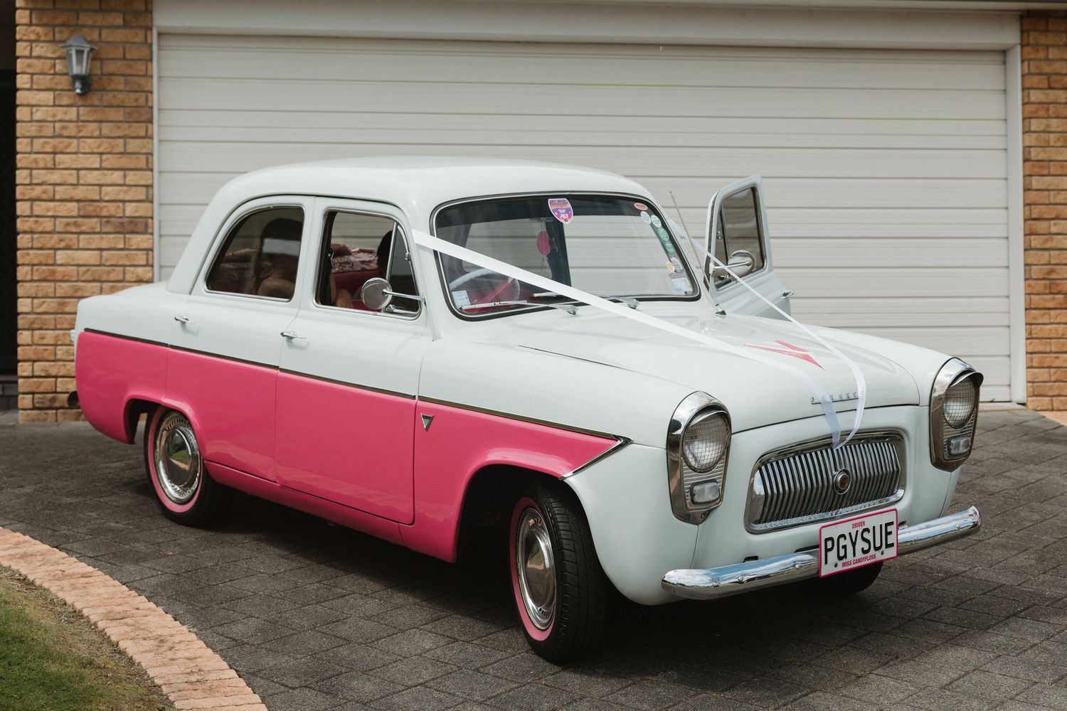 Classic 1950s pink and white Nash Metropolitan car shown from multiple angles on a driveway against brick wall.