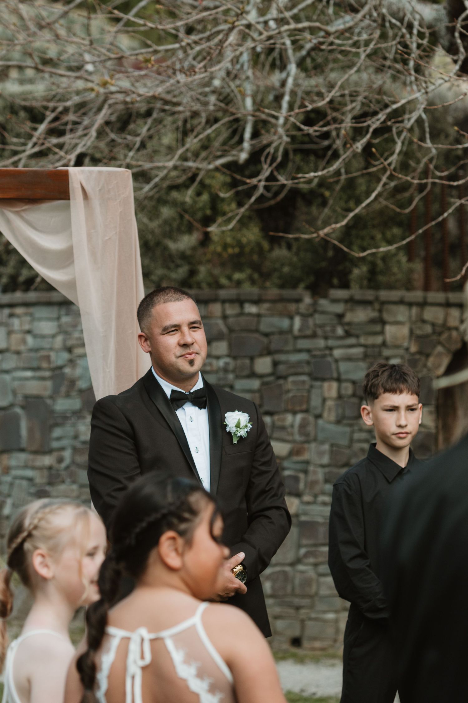 Wedding party members in formal attire stand together during an outdoor ceremony against a stone wall backdrop.