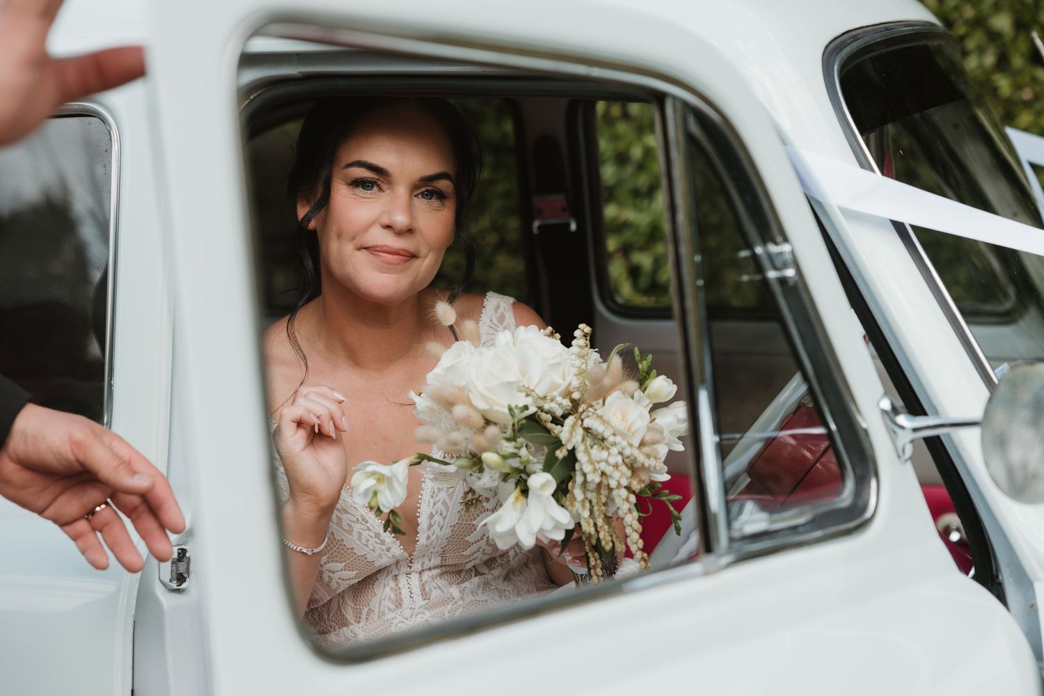 Bride and wedding party members pose through vintage car windows holding white floral bouquets.