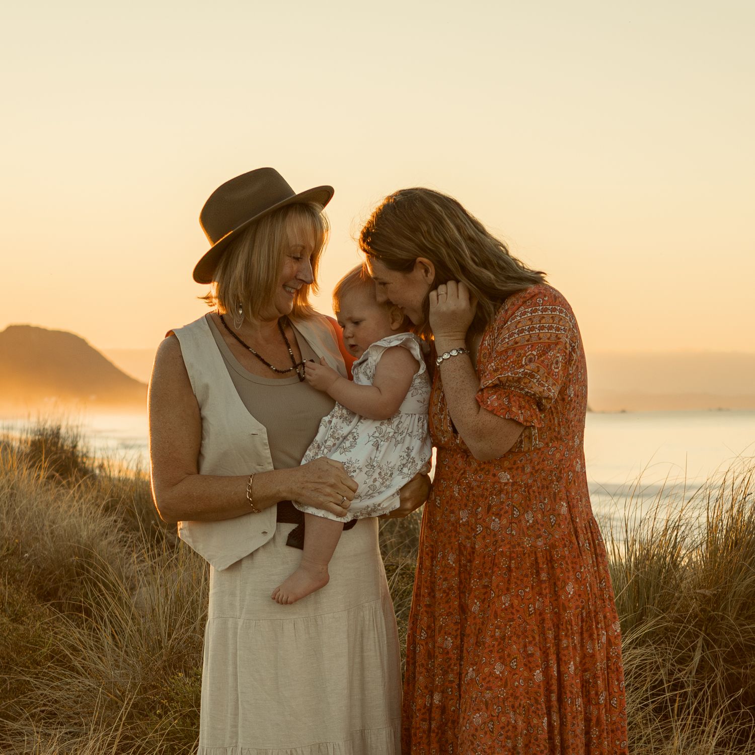 A tender family moment at sunset on the beach with golden light illuminating the coastal grass and ocean backdrop.