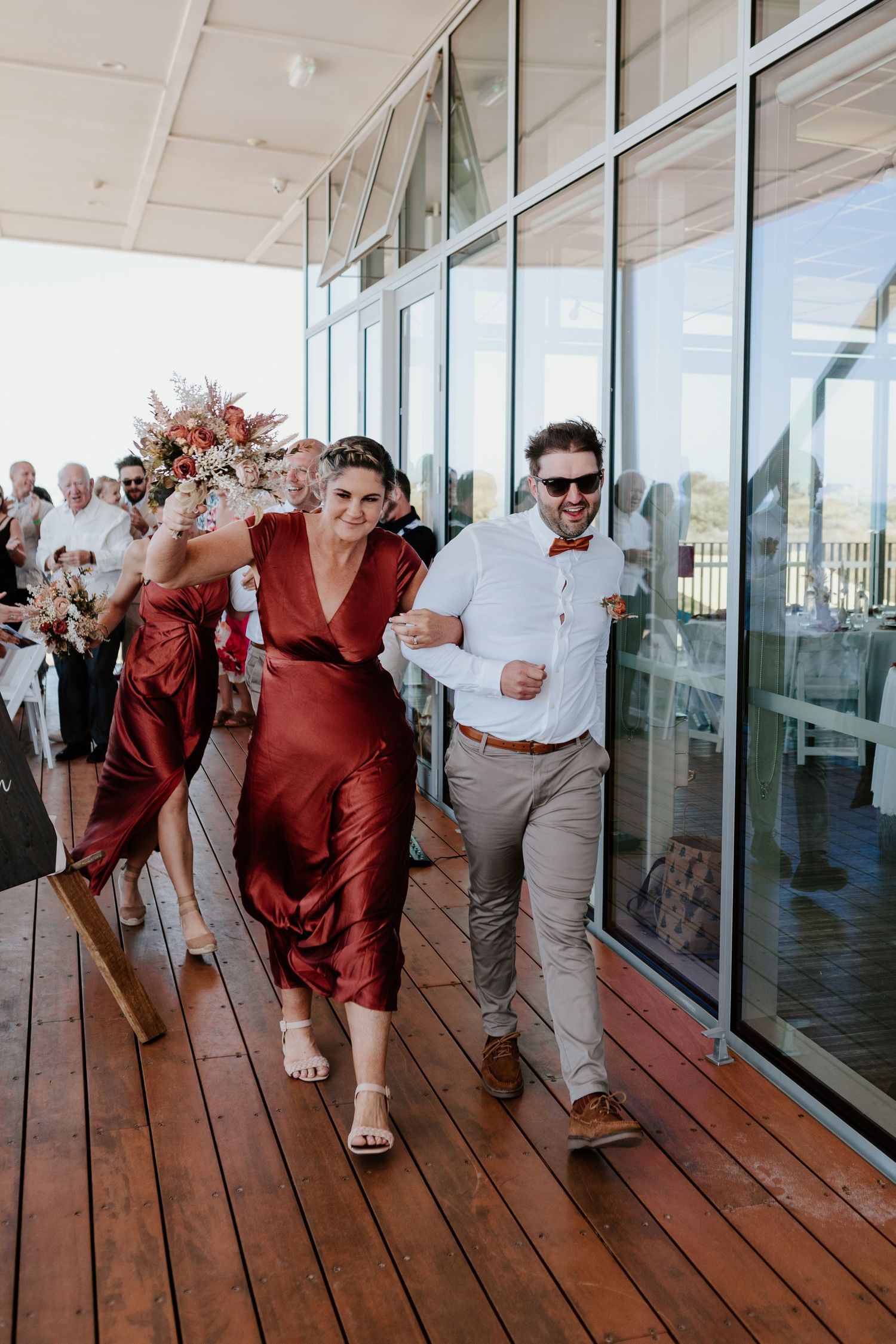 A couple walks joyfully along a wooden deck with glass windows and railings while wearing maroon dress and beige attire.