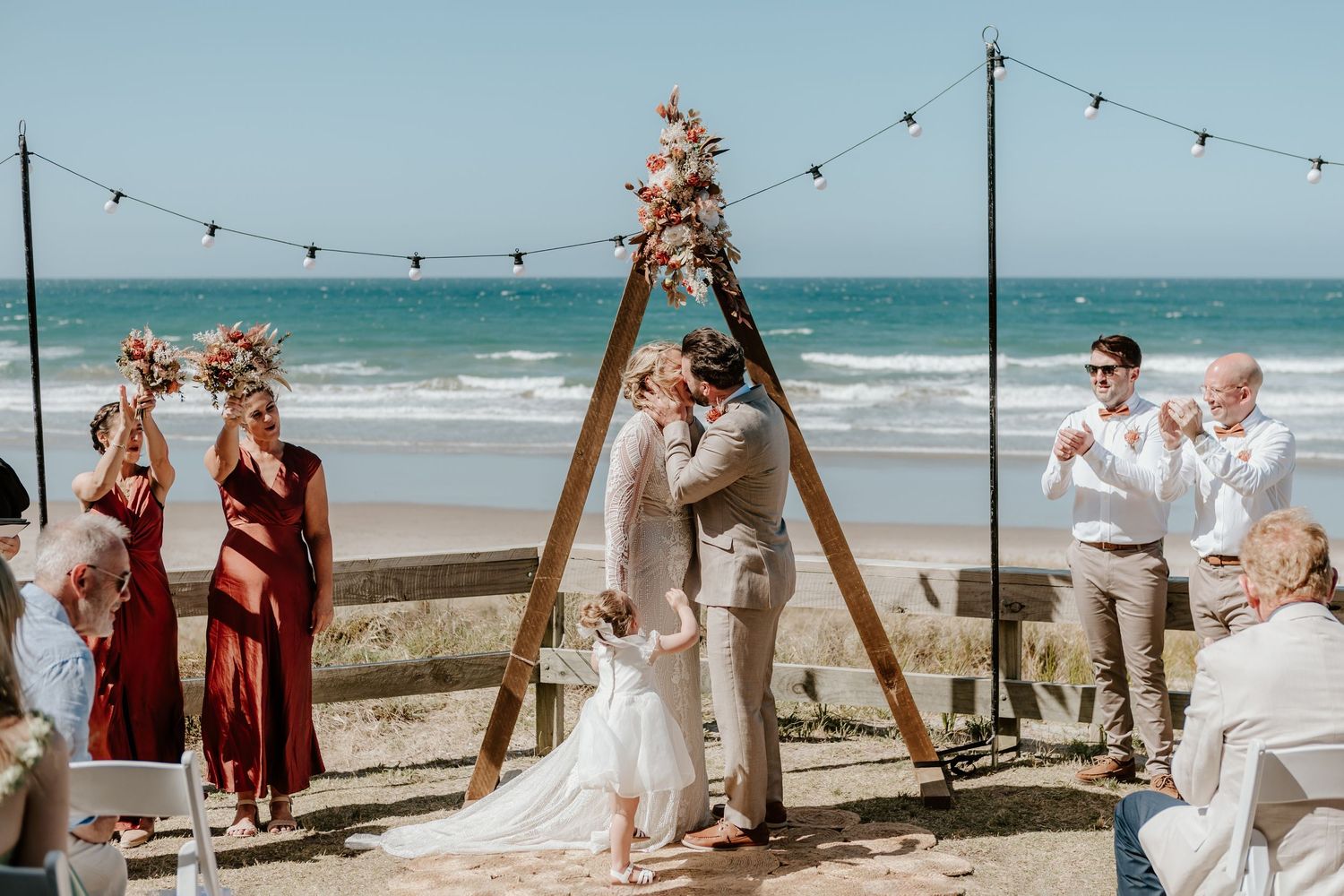 A beachfront wedding ceremony with guests seated in white chairs under string lights beside the ocean.