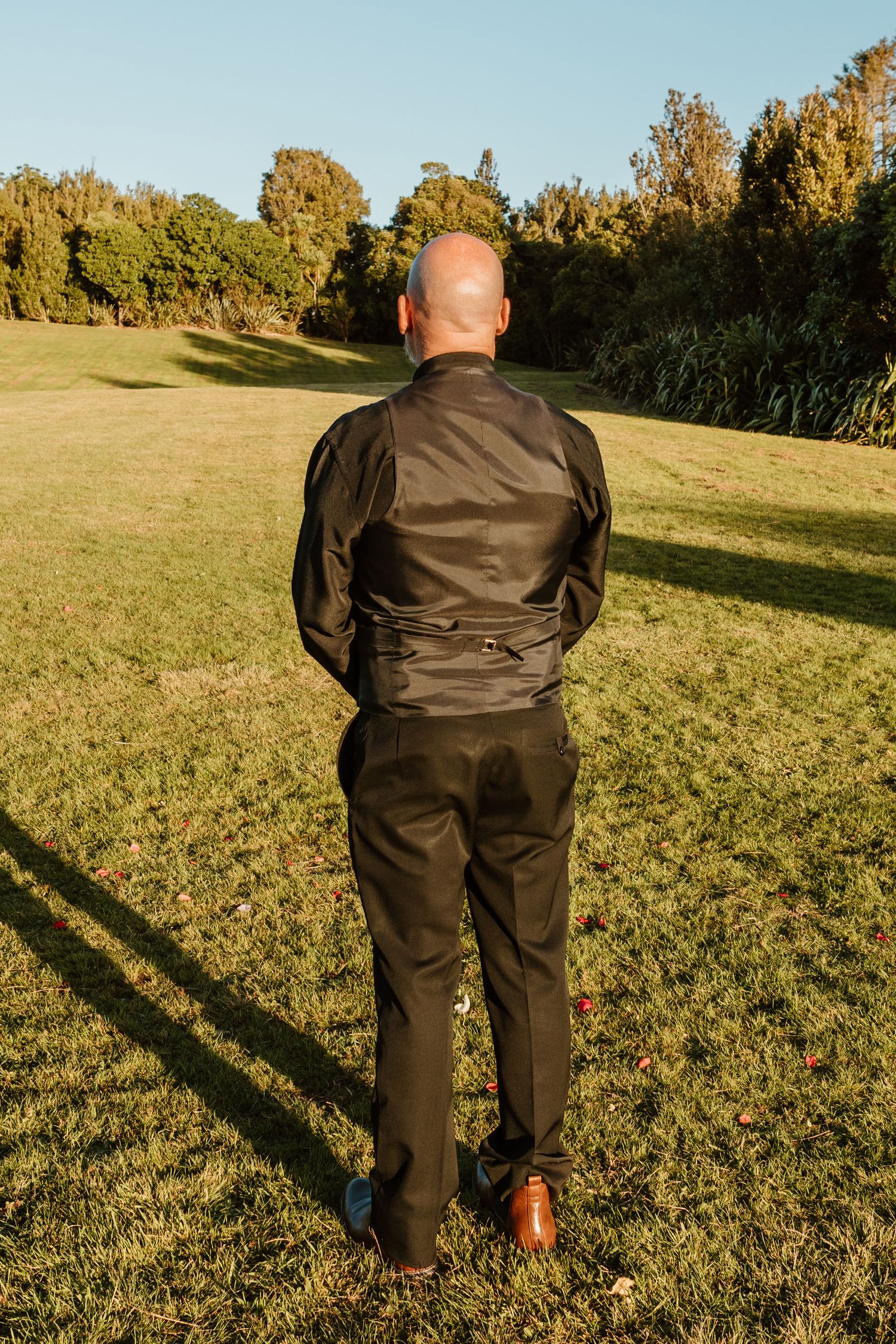 Person wearing brown leather jacket and pants standing in a sunny grassy field with trees in the background.