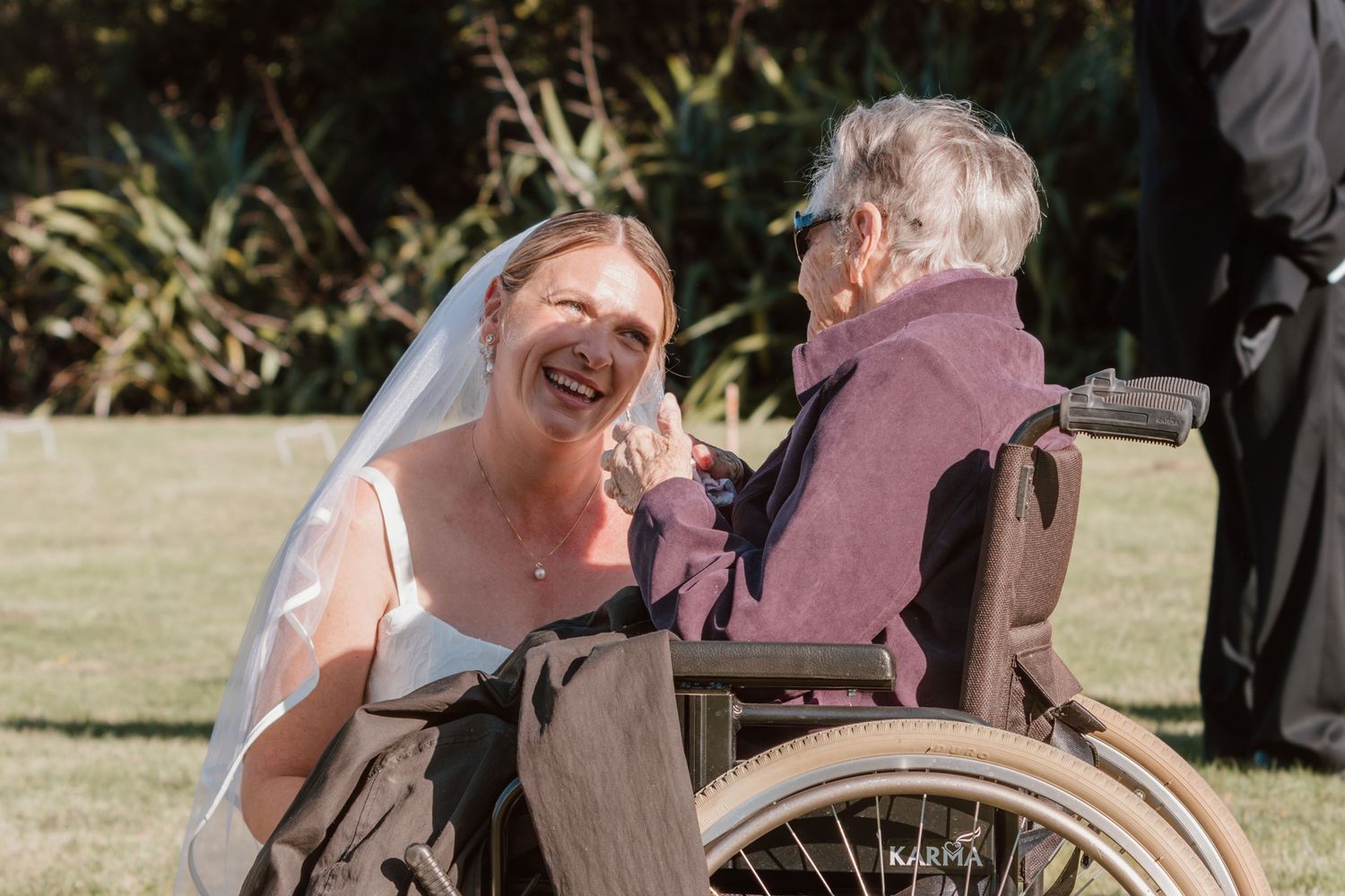 A joyful bride in a white dress shares a heartwarming moment with an elderly person in a wheelchair outdoors.