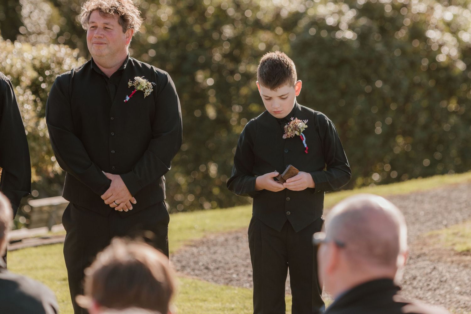 Two people in black attire stand outdoors during what appears to be a formal ceremony or gathering on a sunny day.