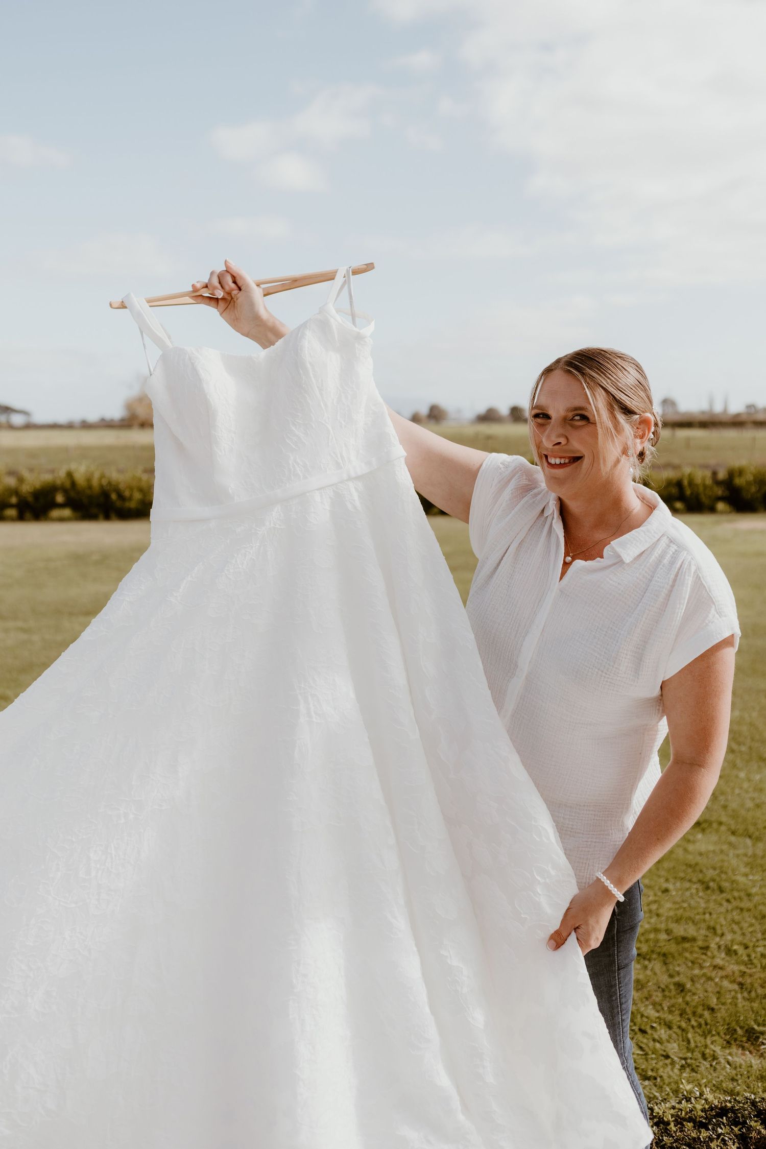 Woman in white shirt holds up an elegant white wedding dress on a sunny day in a grassy outdoor setting.