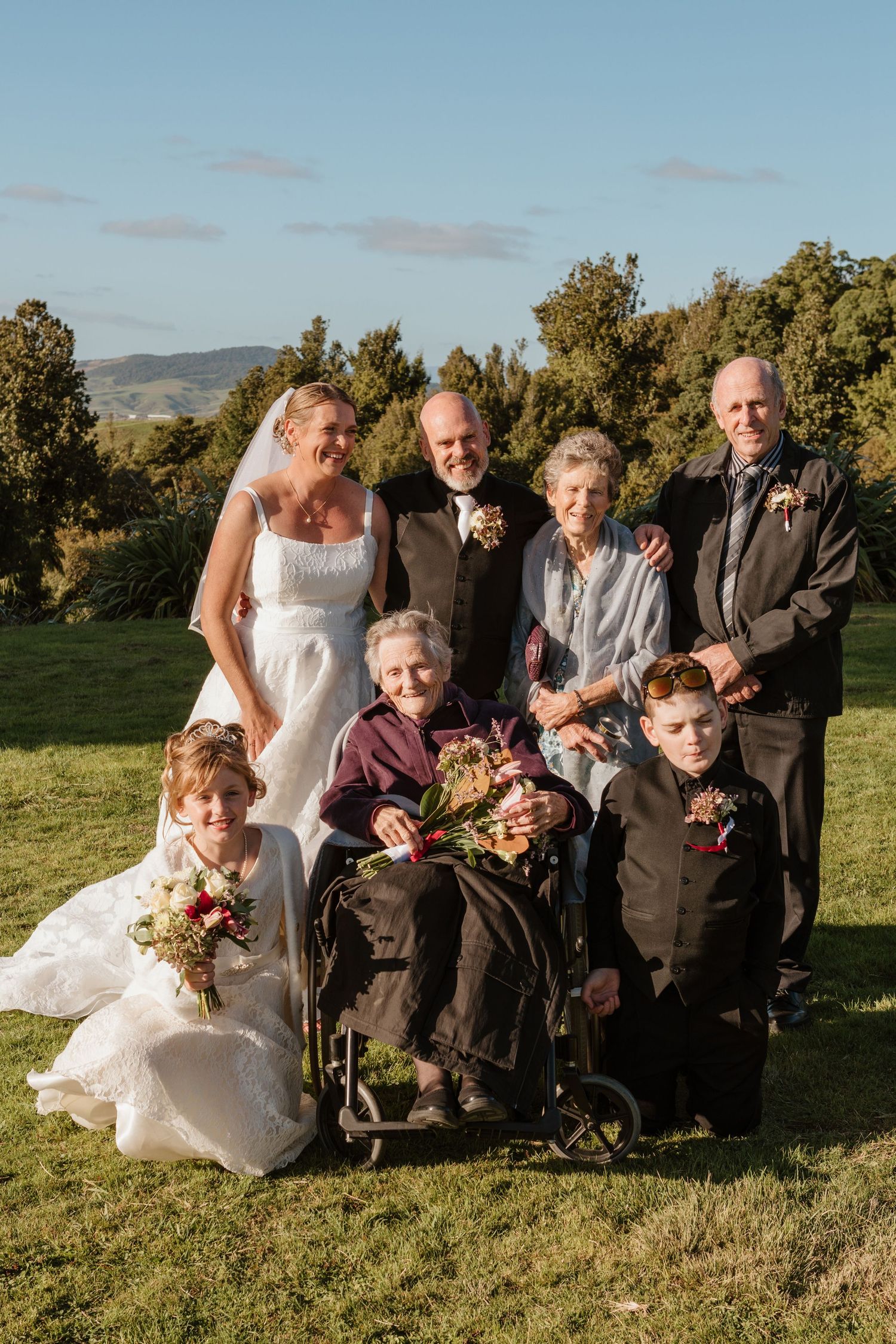 Wedding party poses together outdoors on grassy hillside with mountains and blue sky in background.