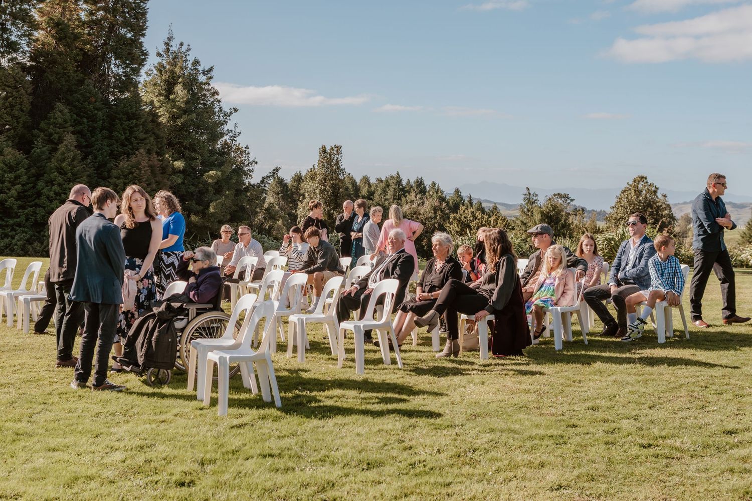A group of wedding guests seated on white chairs on a grassy lawn during an outdoor ceremony.