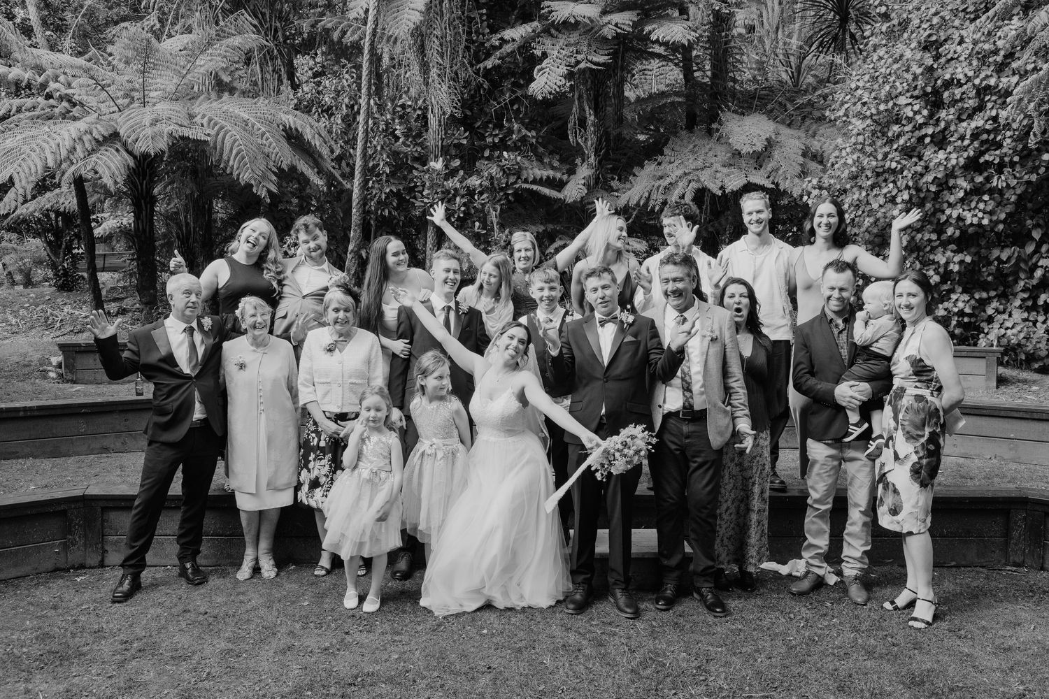 A black and white wedding photo of a large group gathered outdoors with tropical palm trees in the background.