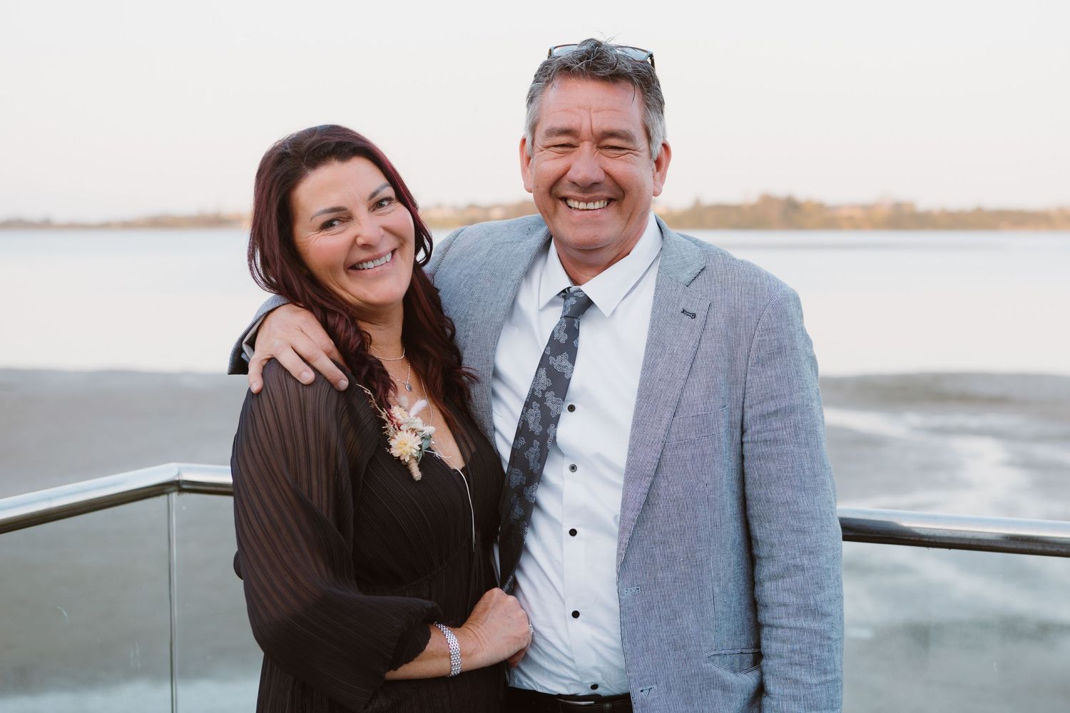 A couple smiles together while standing on a rooftop deck overlooking a waterfront view at sunset.