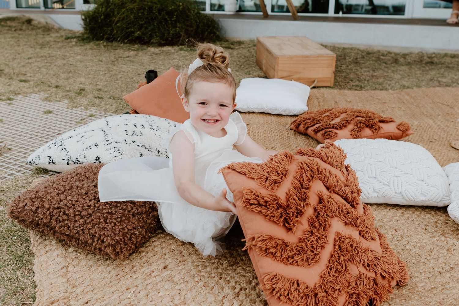 A small child sits among white pillows and terracotta blankets at a beach wedding setting. Papamoa Surf Lifesaving Club Wedding