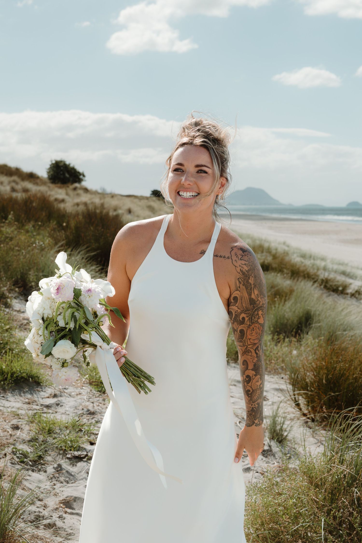 Smiling bride in white halter dress with floral tattoo holds bouquet on sandy beach with blue sky backdrop.