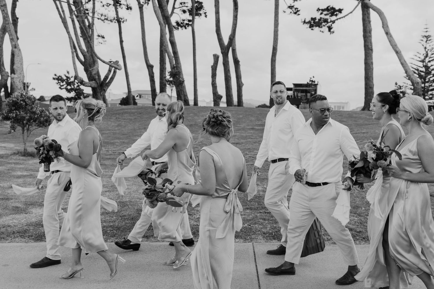 Black and white photo of a wedding party walking outdoors near trees, bridesmaids in elegant gowns and groomsmen in white attire.
