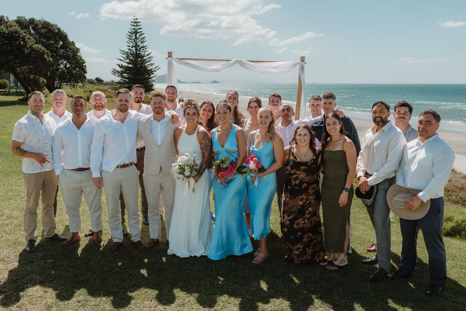 Wedding party poses together on a grassy beachfront with an arch and ocean backdrop on a sunny day.