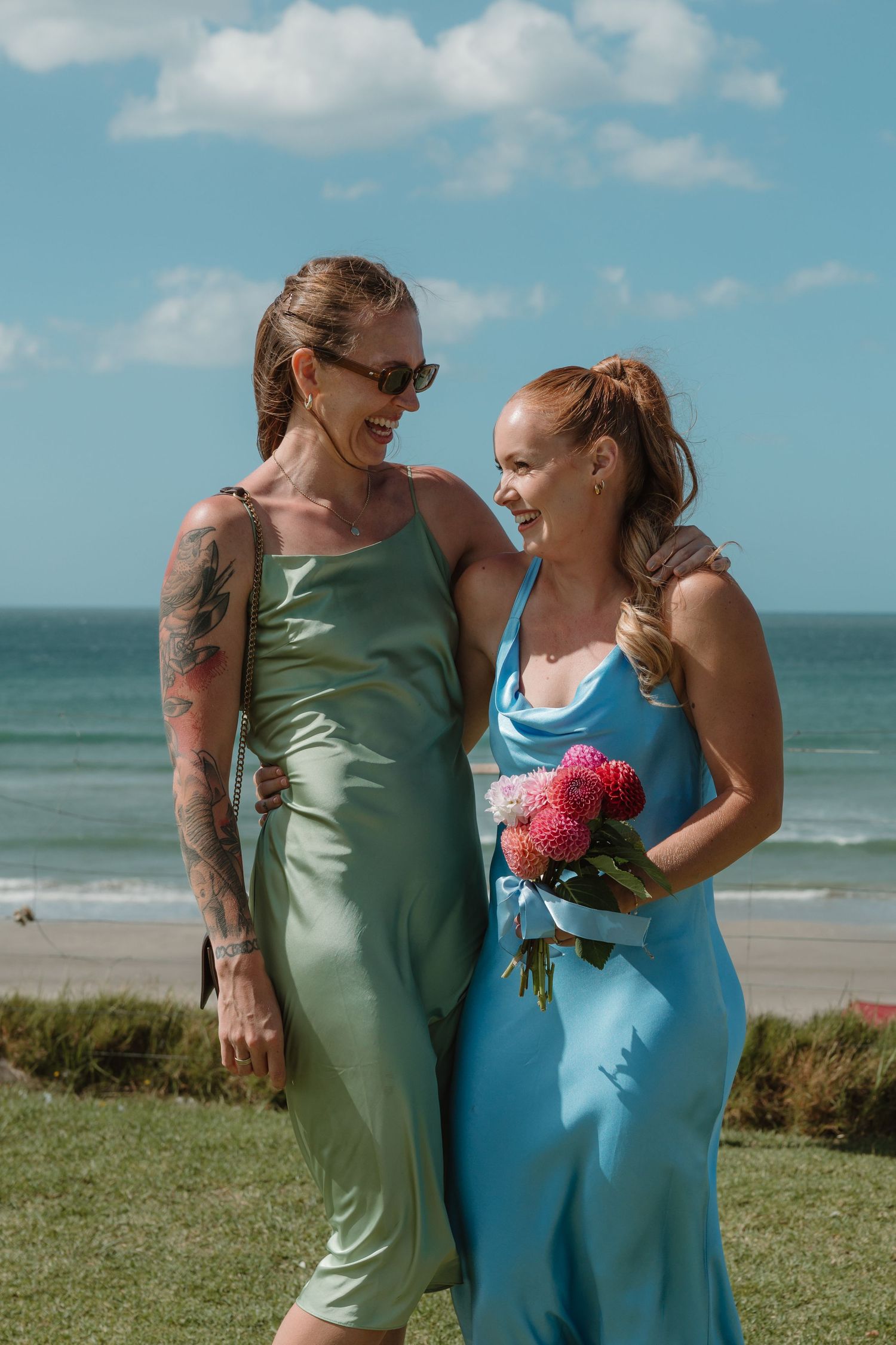 Two women in elegant satin slip dresses smile at each other on a beachside lawn, one holding a pink floral bouquet.