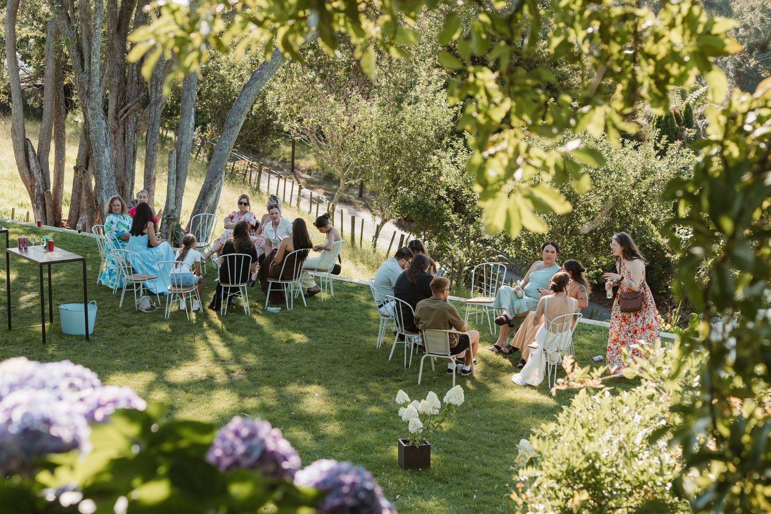 Guests seated on white chairs in a lush garden with hydrangeas and trees during an outdoor event.