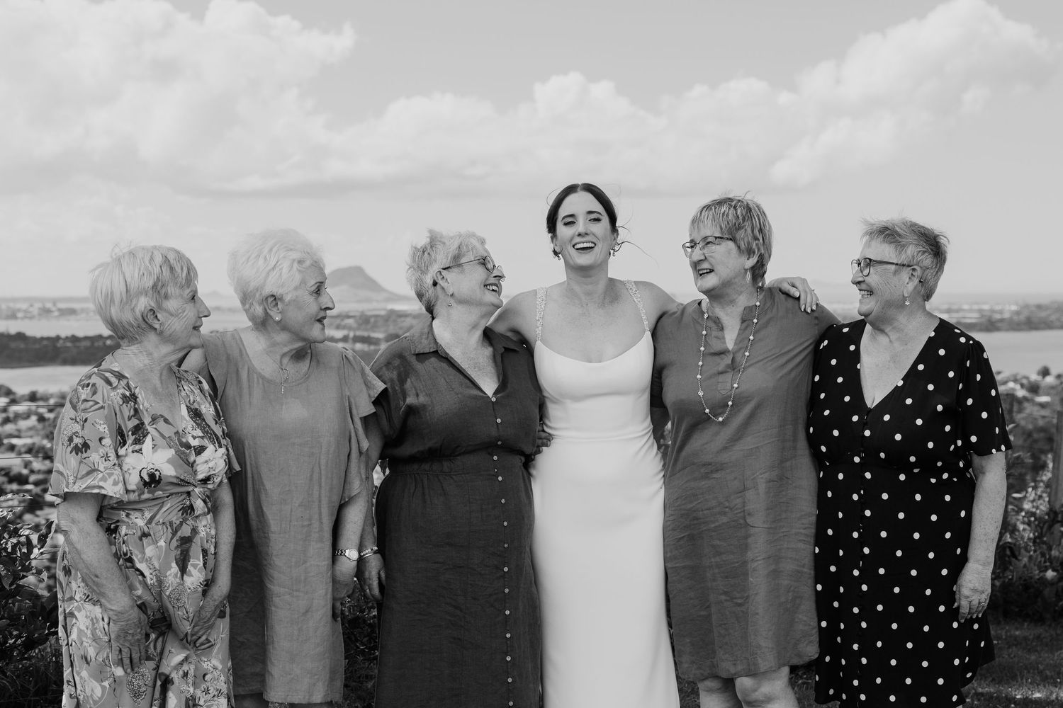 Bride in white dress laughing with five women outdoors in black and white wedding photo.