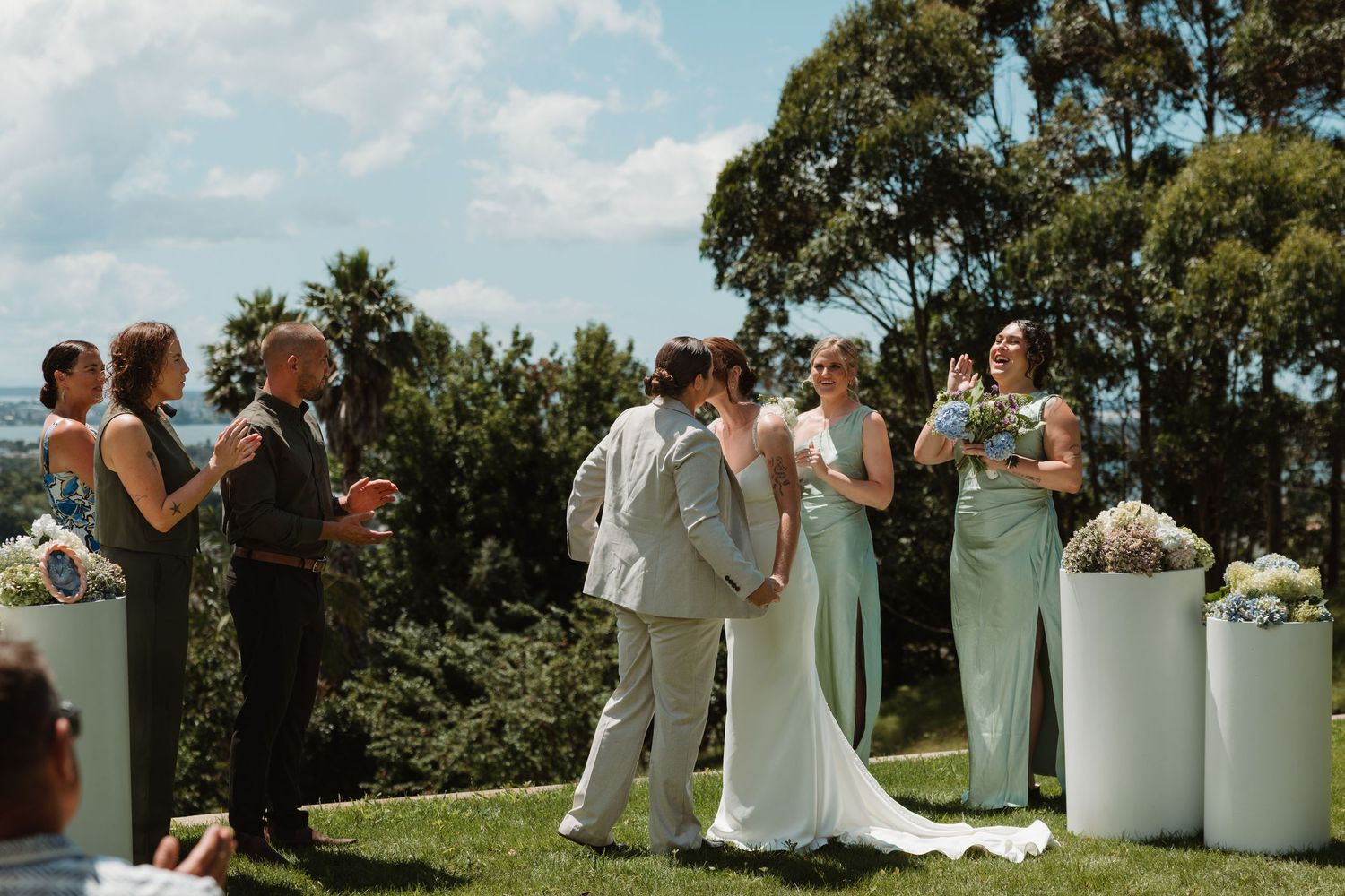 Bride and groom share first kiss at outdoor wedding ceremony with bridesmaids in sage green dresses.