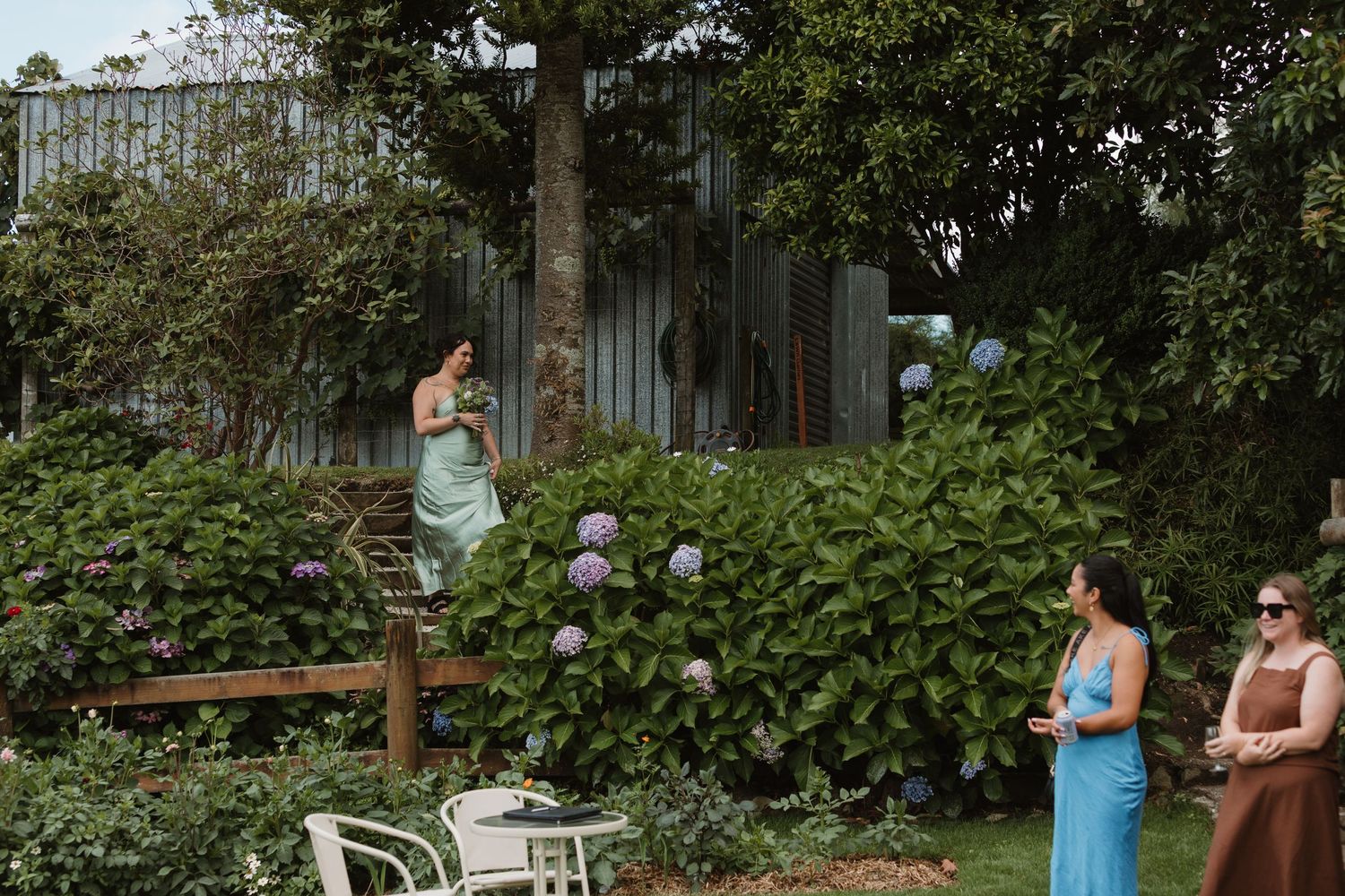 Women in elegant dresses gather in a lush garden with hydrangeas and a rustic corrugated building backdrop.