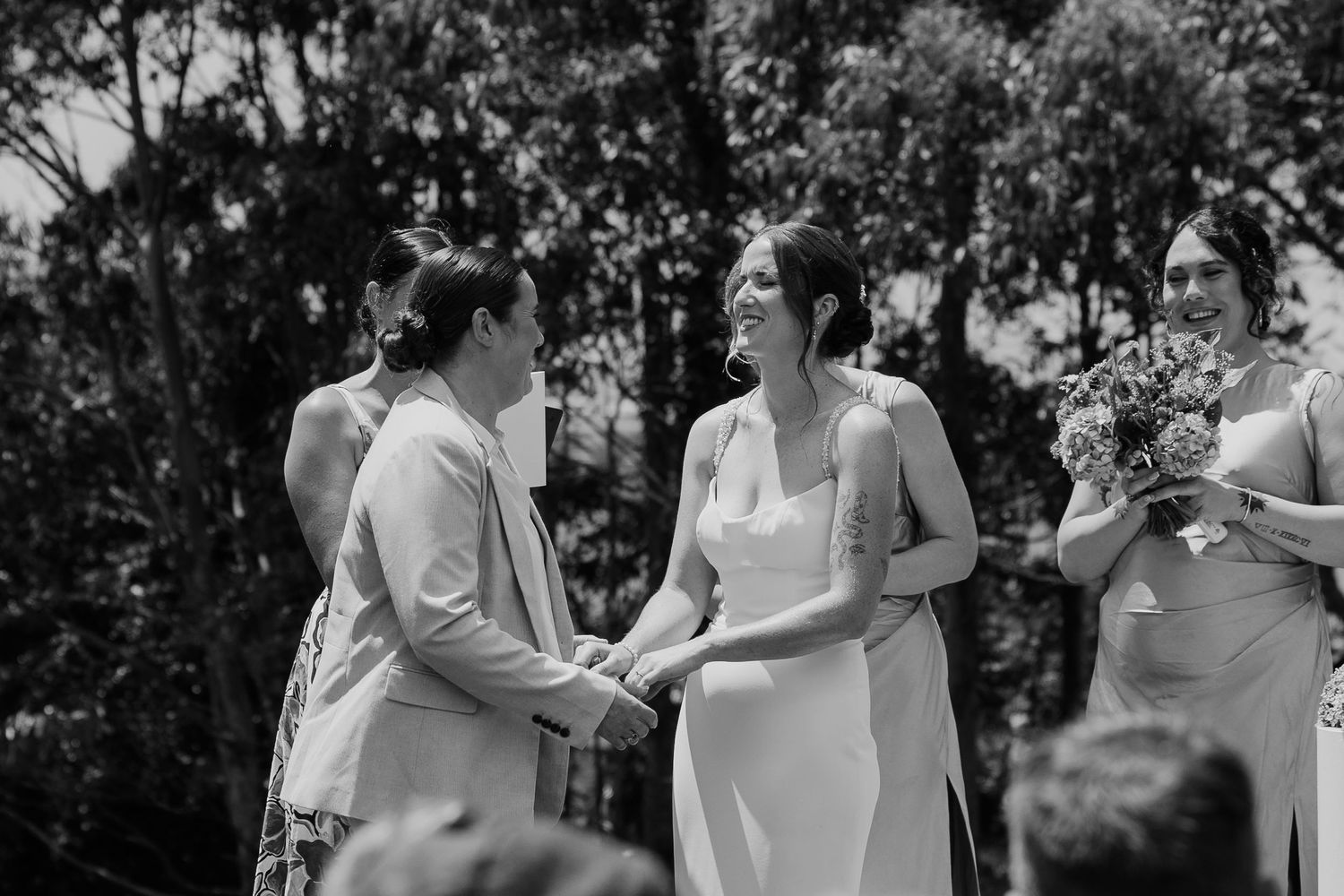 Black and white photo of two brides holding hands and smiling during outdoor wedding ceremony with bridesmaid holding bouquet.