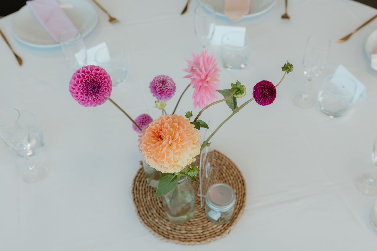 Colorful dahlia flowers in a glass jar vase on a wicker mat, wedding table centerpiece with pink and orange blooms.