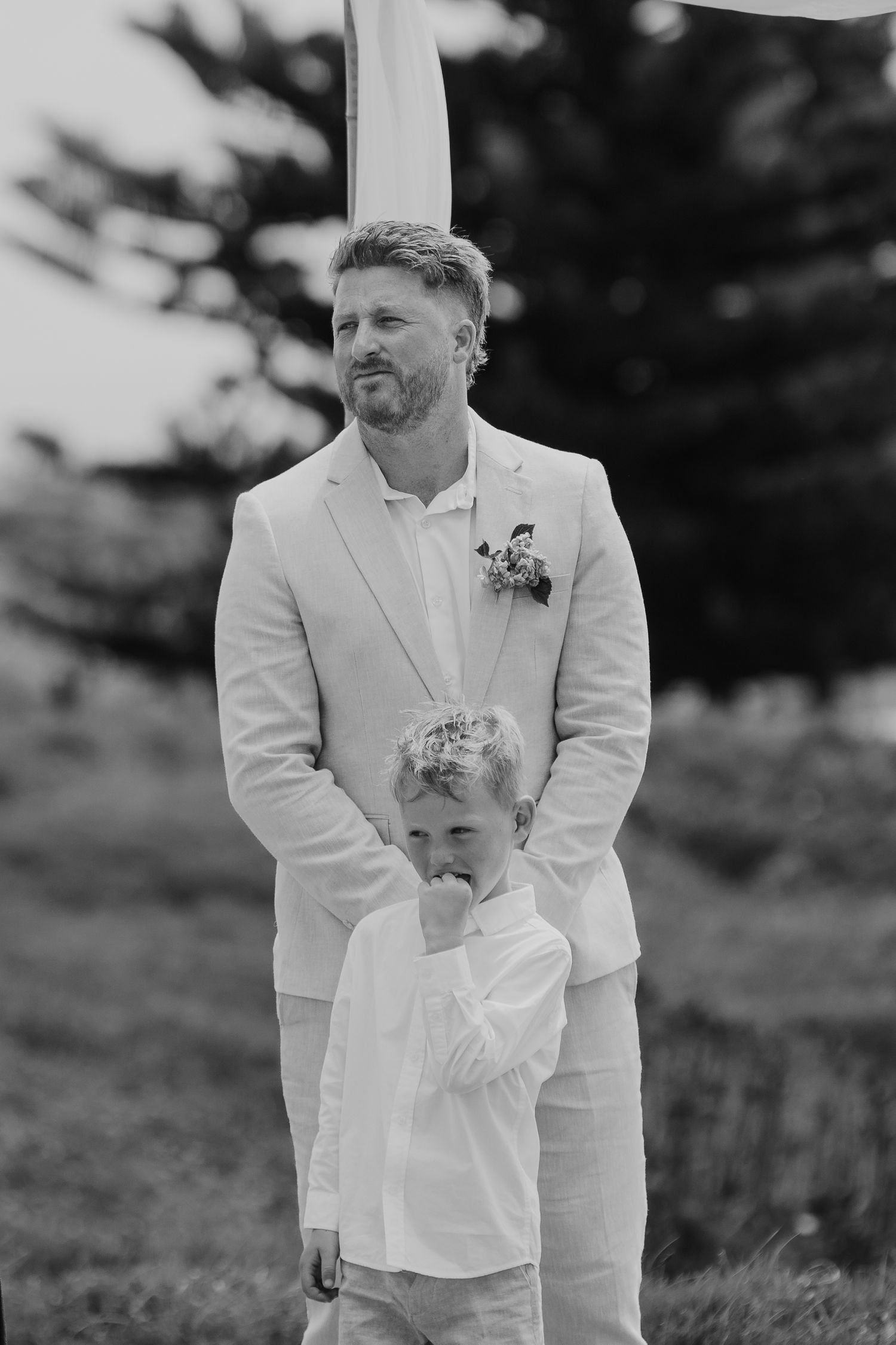 Black and white photo of a groom in a white suit with boutonniere standing behind a young boy at an outdoor wedding.