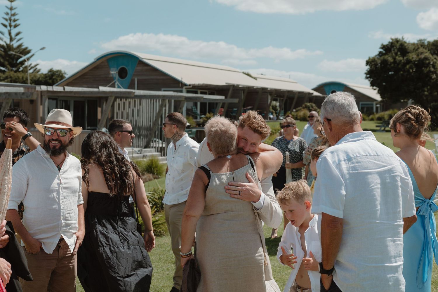 Guests mingling outdoors at a coastal wedding venue on a sunny day, dressed in smart casual attire.
