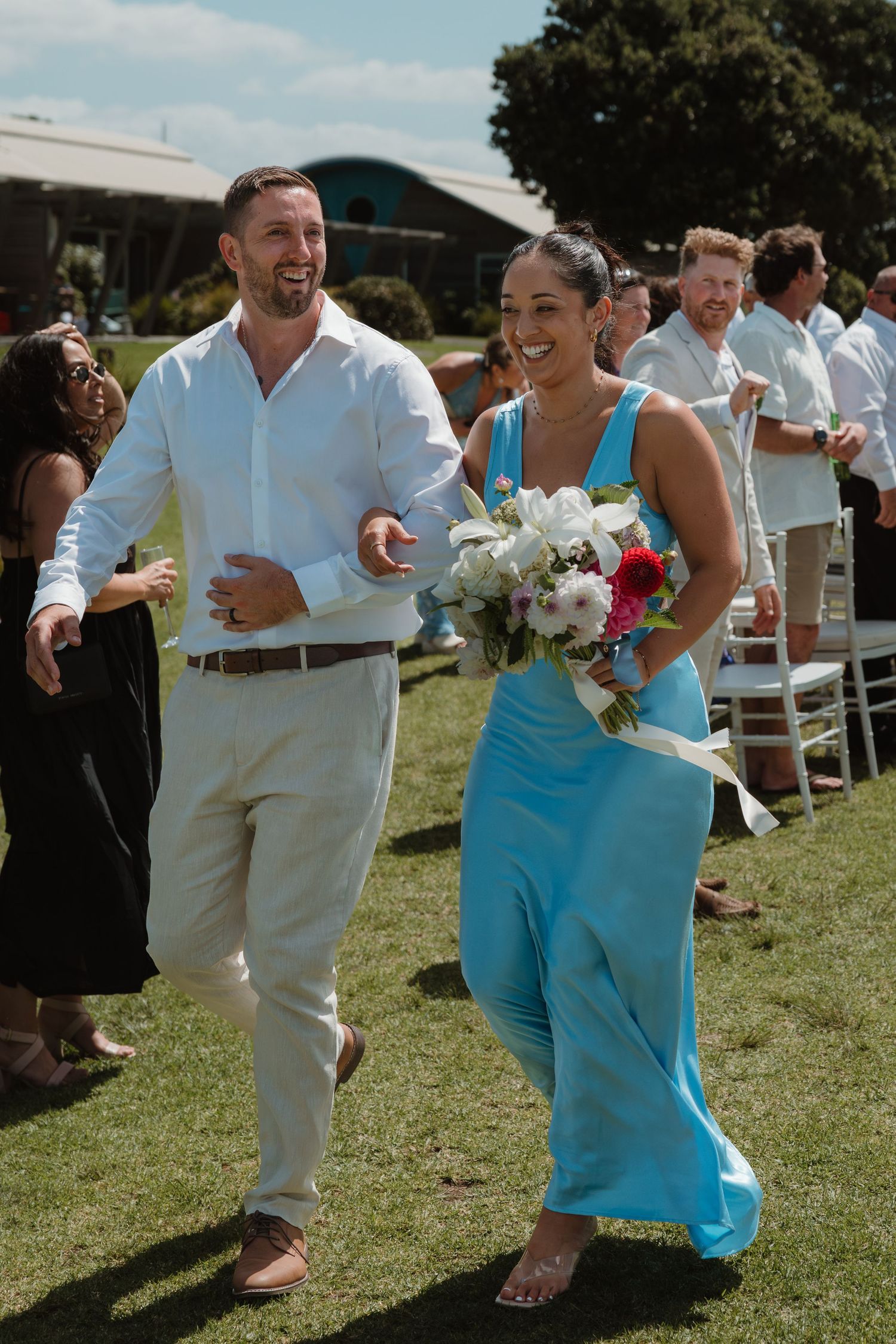 Bridesmaid in turquoise dress walks down outdoor wedding aisle carrying white and pink bouquet beside groomsman in white shirt.