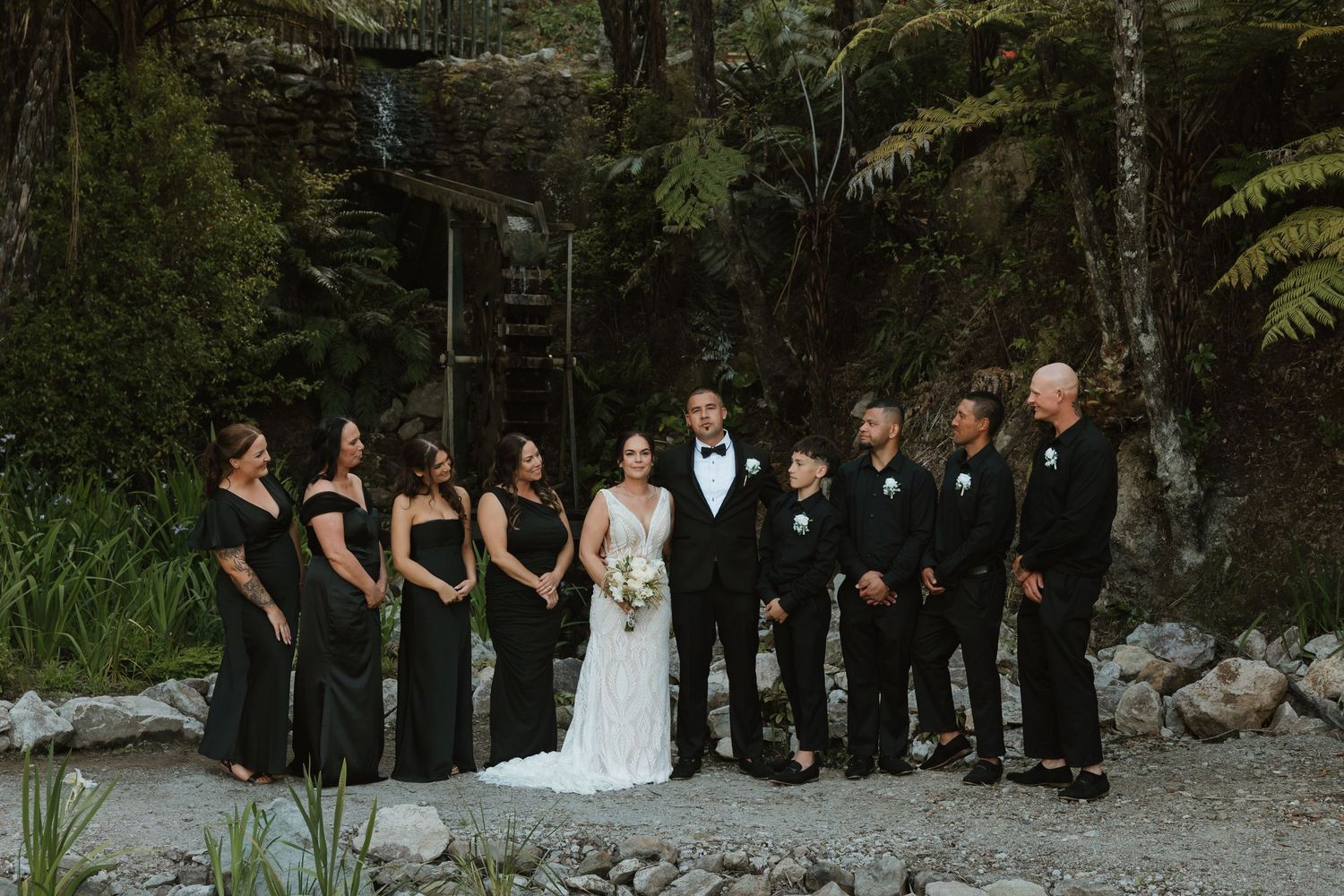 Wedding party members stand in a row outdoors against a lush forest backdrop during a formal ceremony.