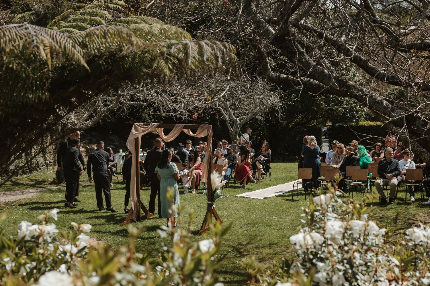 Wedding guests gather outdoors on a sunny day surrounded by lush greenery and white floral decorations.