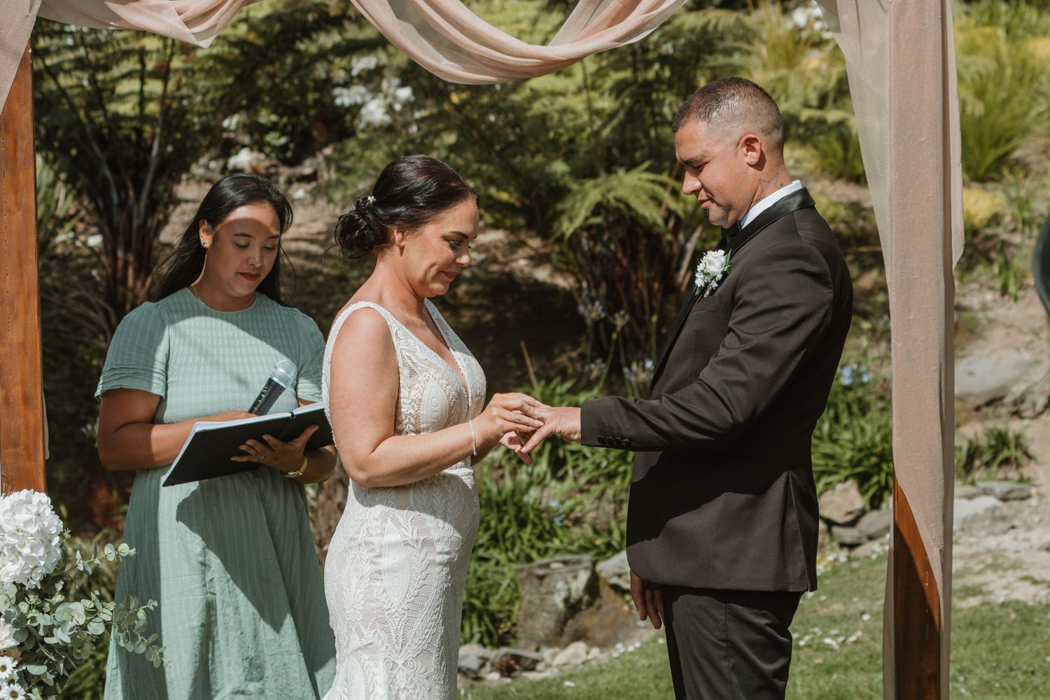 Wedding party stands under a rustic wooden pergola during an outdoor ceremony in a lush garden setting.