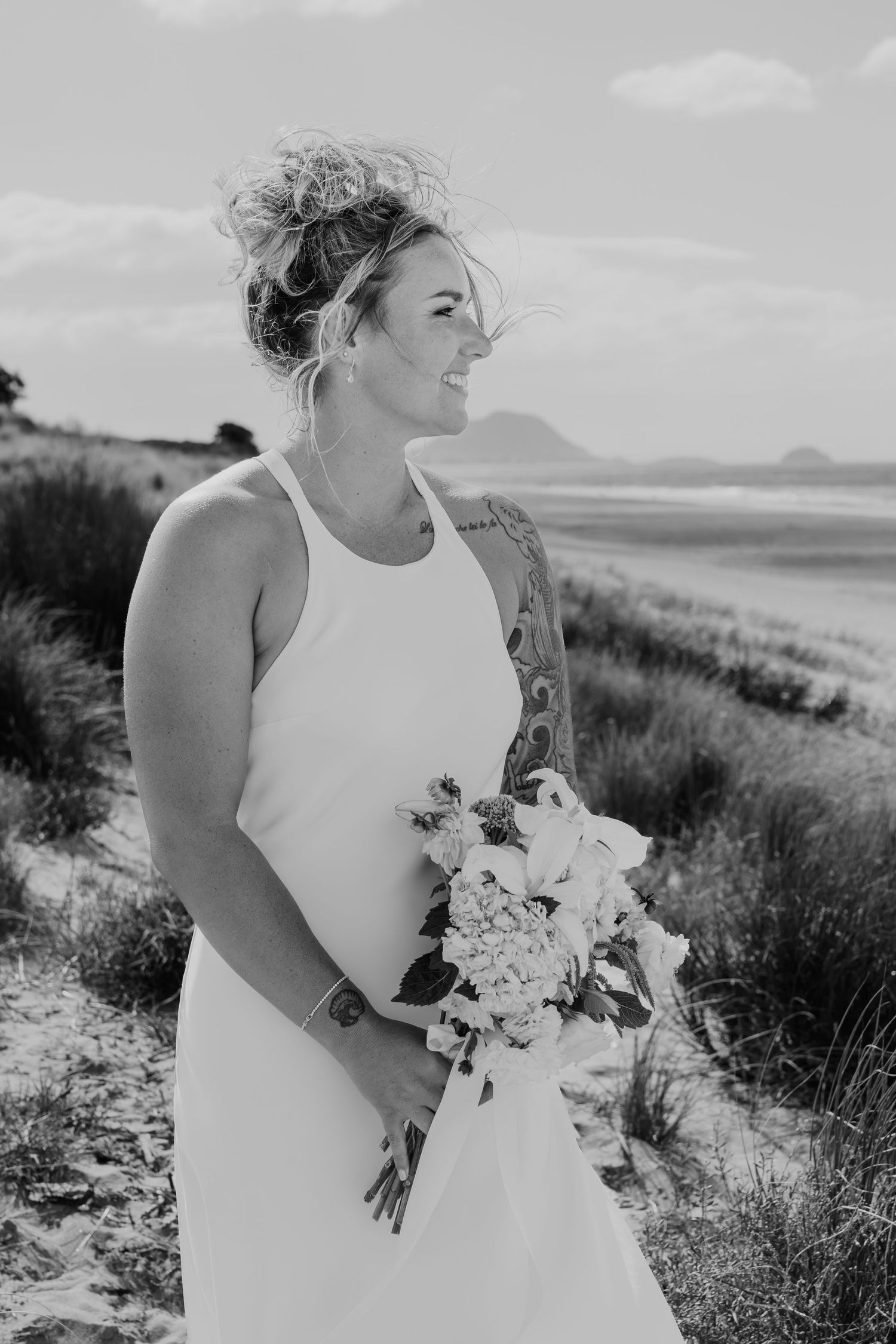 Black and white photo of a bride in a white dress holding a bouquet on a sandy beach with ocean views.