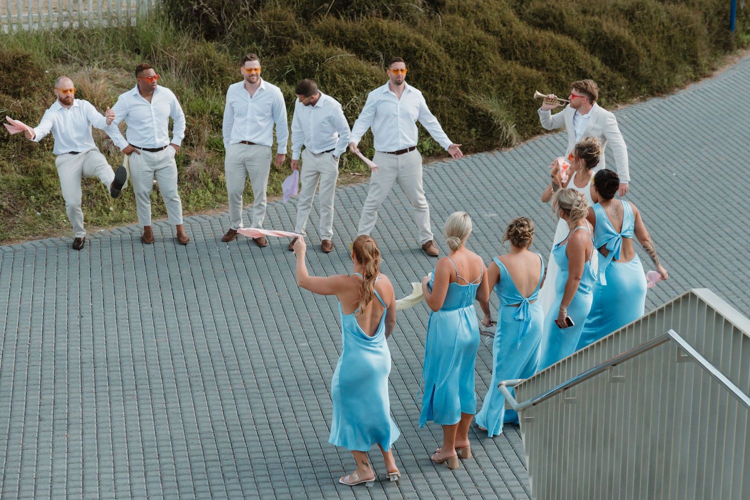 Wedding party in blue dresses and white suits celebrating on a rooftop with greenery backdrop.