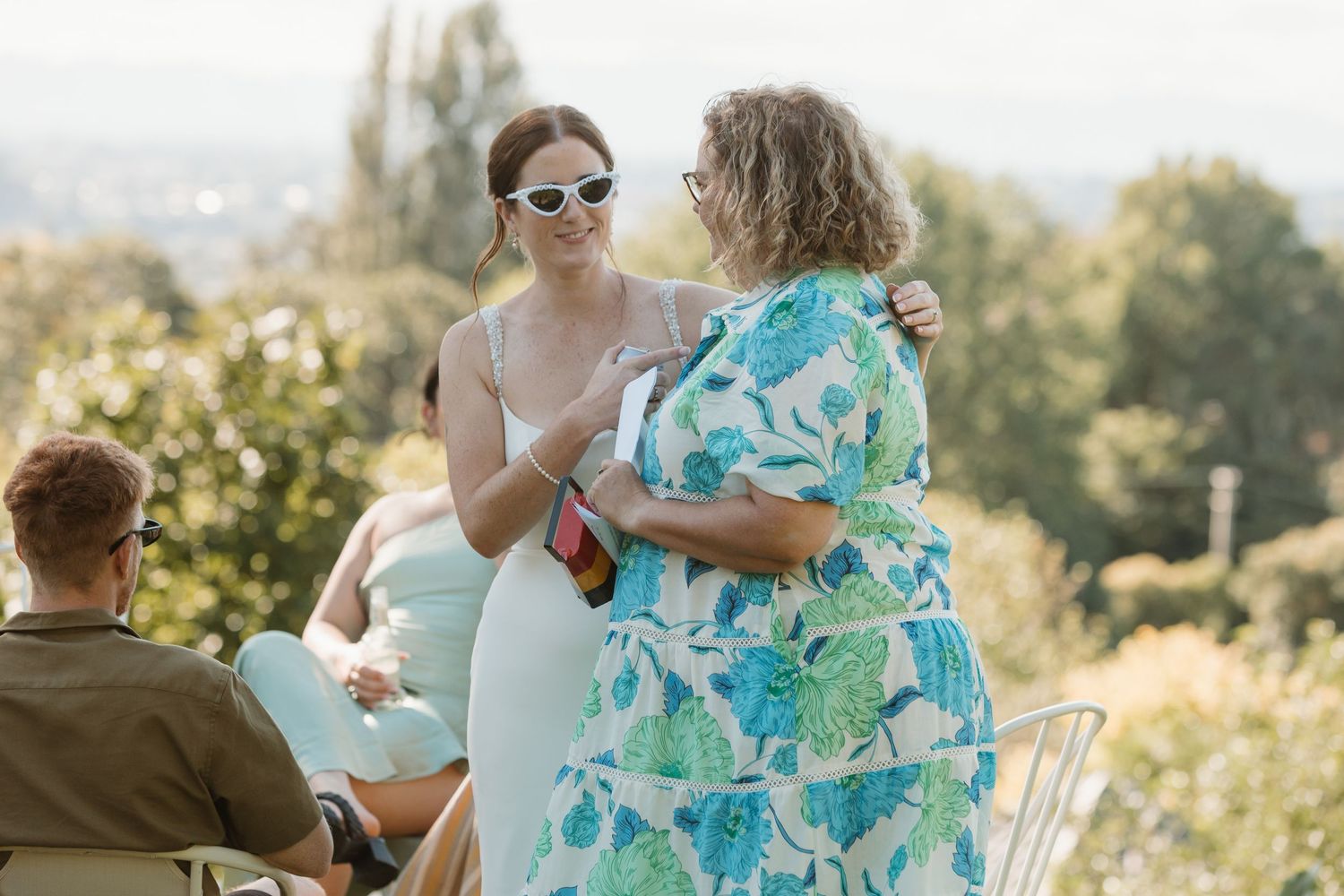 Two women in summer dresses chat outdoors at a garden party with scenic views in the background.