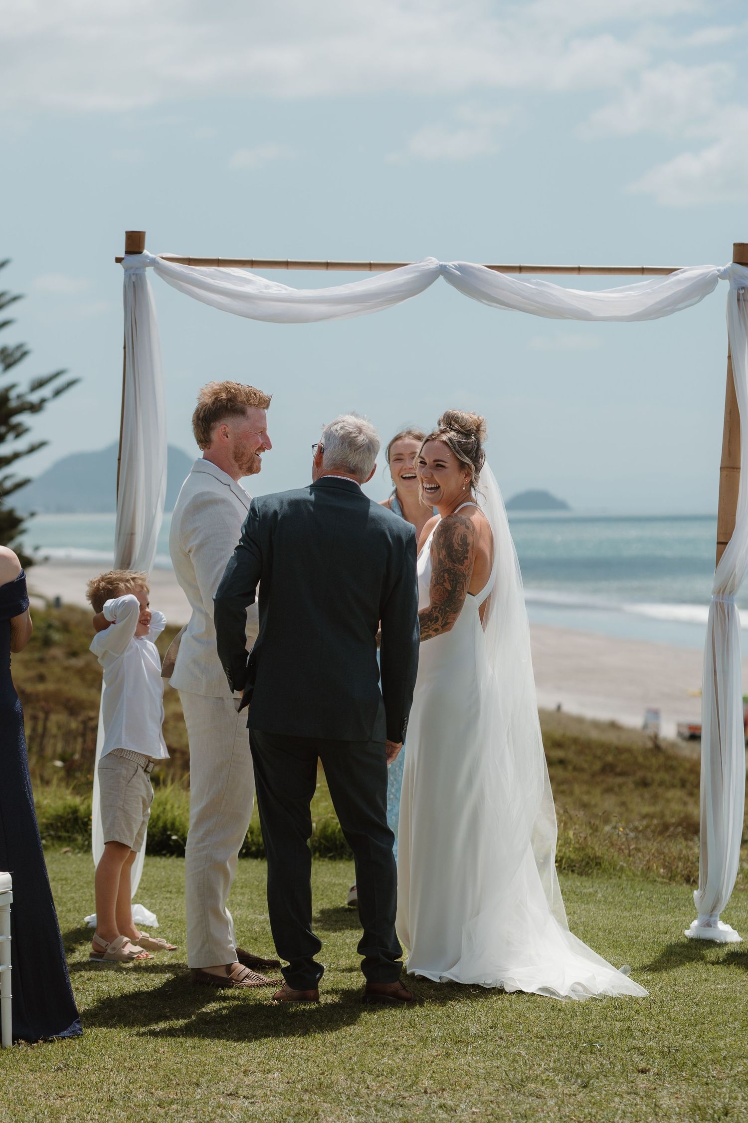 Beach wedding ceremony with bride and groom exchanging vows under a white draped arch overlooking the ocean.