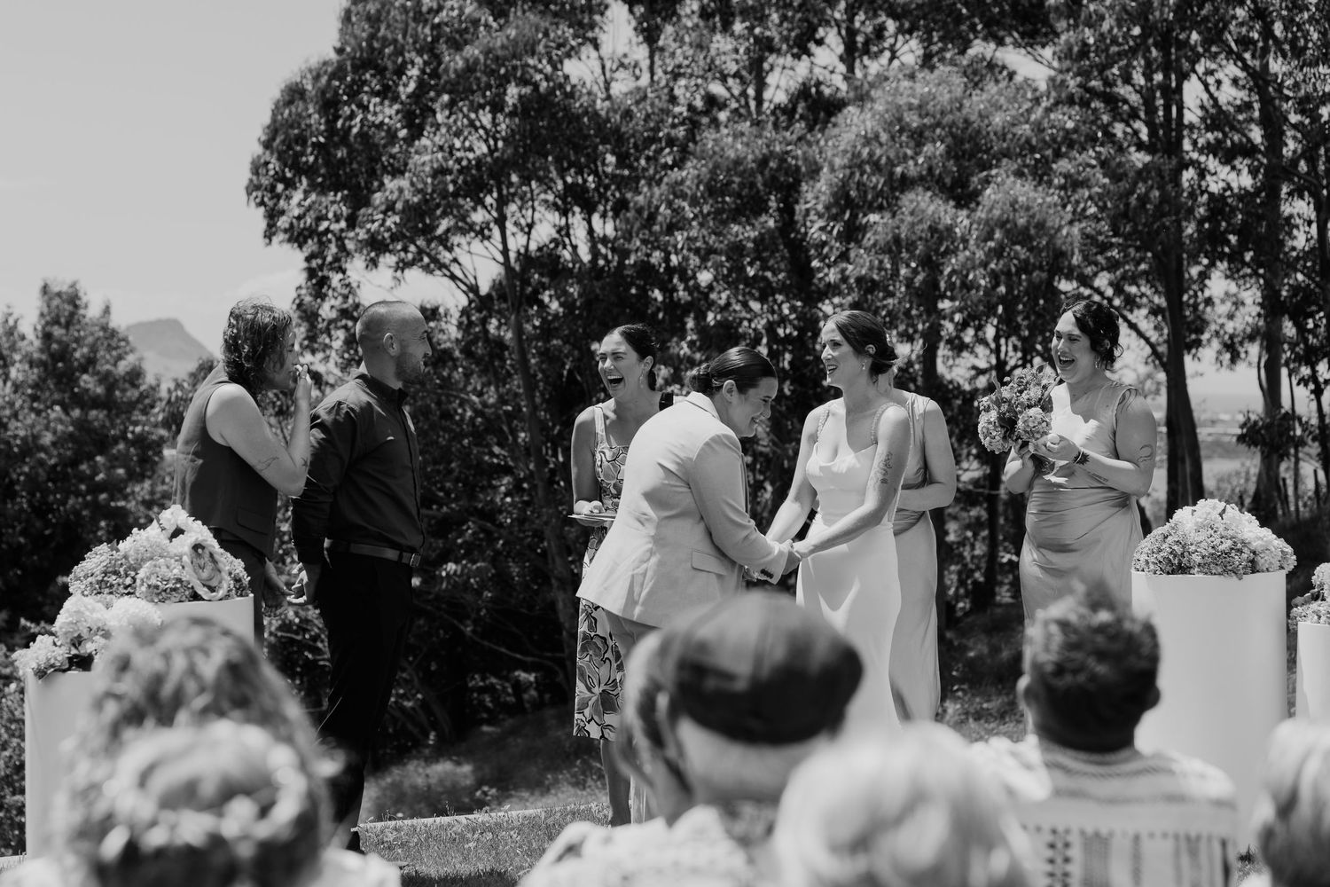 Black and white outdoor wedding ceremony with couple holding hands before officiant, guests seated, surrounded by lush trees.