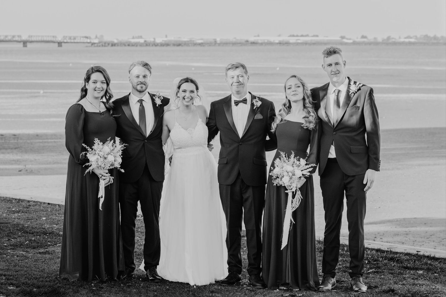 Wedding party members pose together in formal attire against a waterfront backdrop.