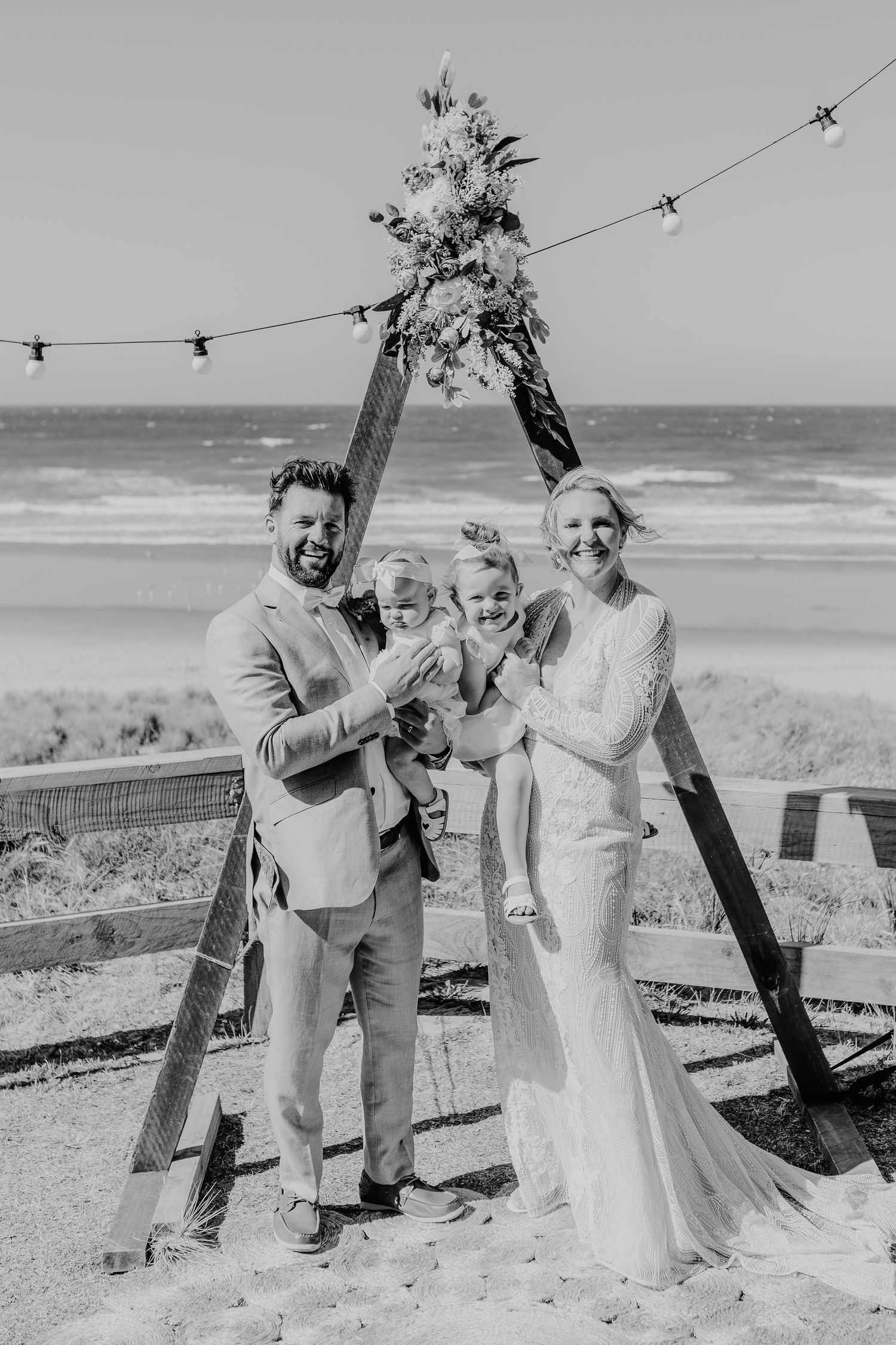 A wedding ceremony moment captured under a wooden arch with string lights and ocean views.  Papamoa Surf Lifesaving Club Wedding