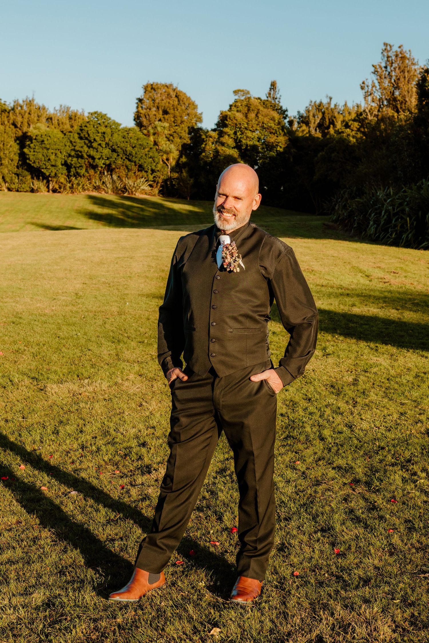 A person in brown business casual attire stands on a sunlit lawn with autumn trees in the background at golden hour.