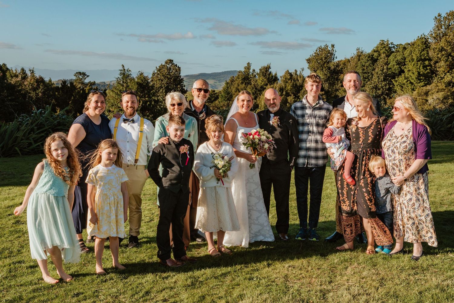 A group dressed in formal and casual attire stands together on a grassy hillside during golden hour.